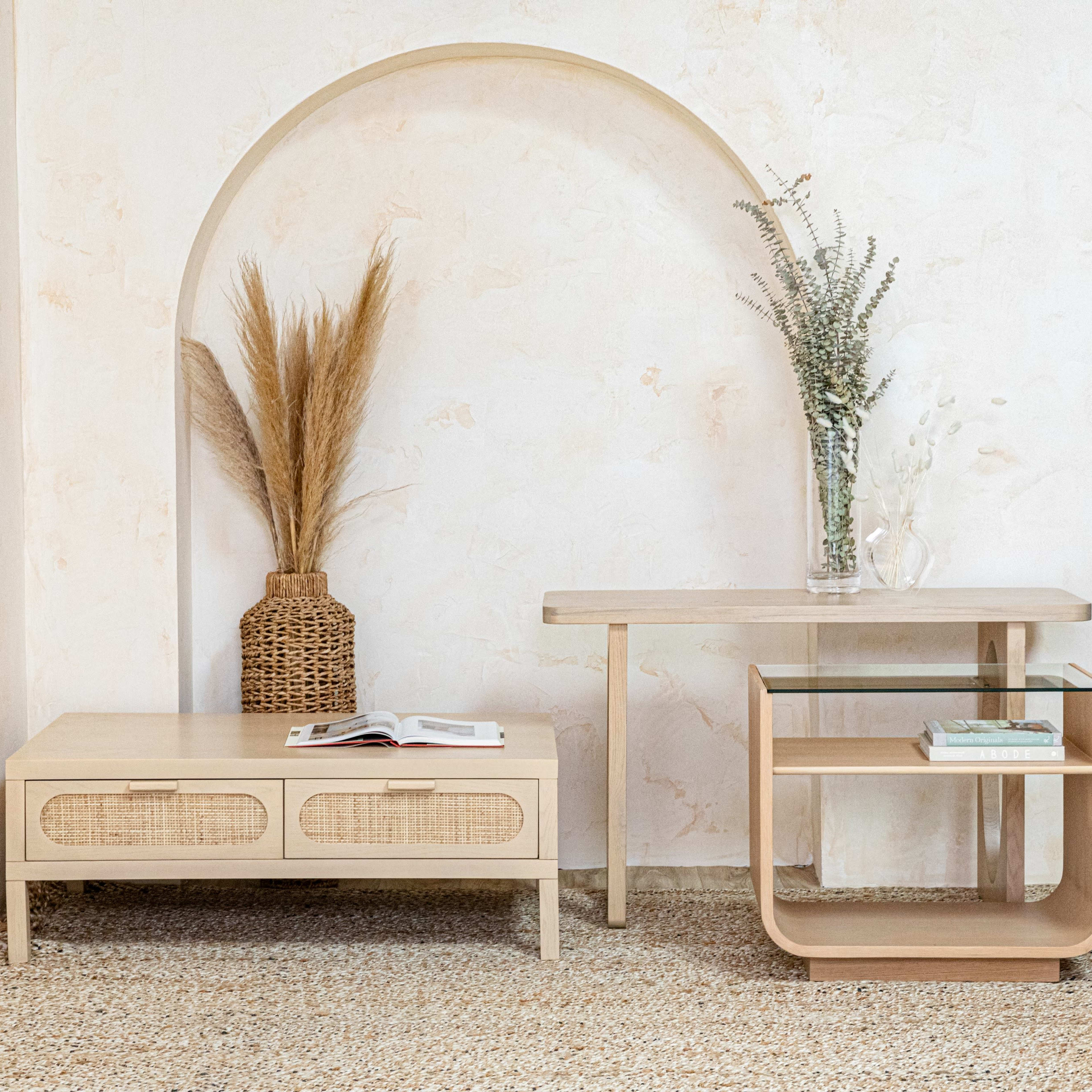 A light-colored room features a minimalist setup with a wooden table, a woven basket holding pampas grass, and a shelf, all positioned on a textured rug.