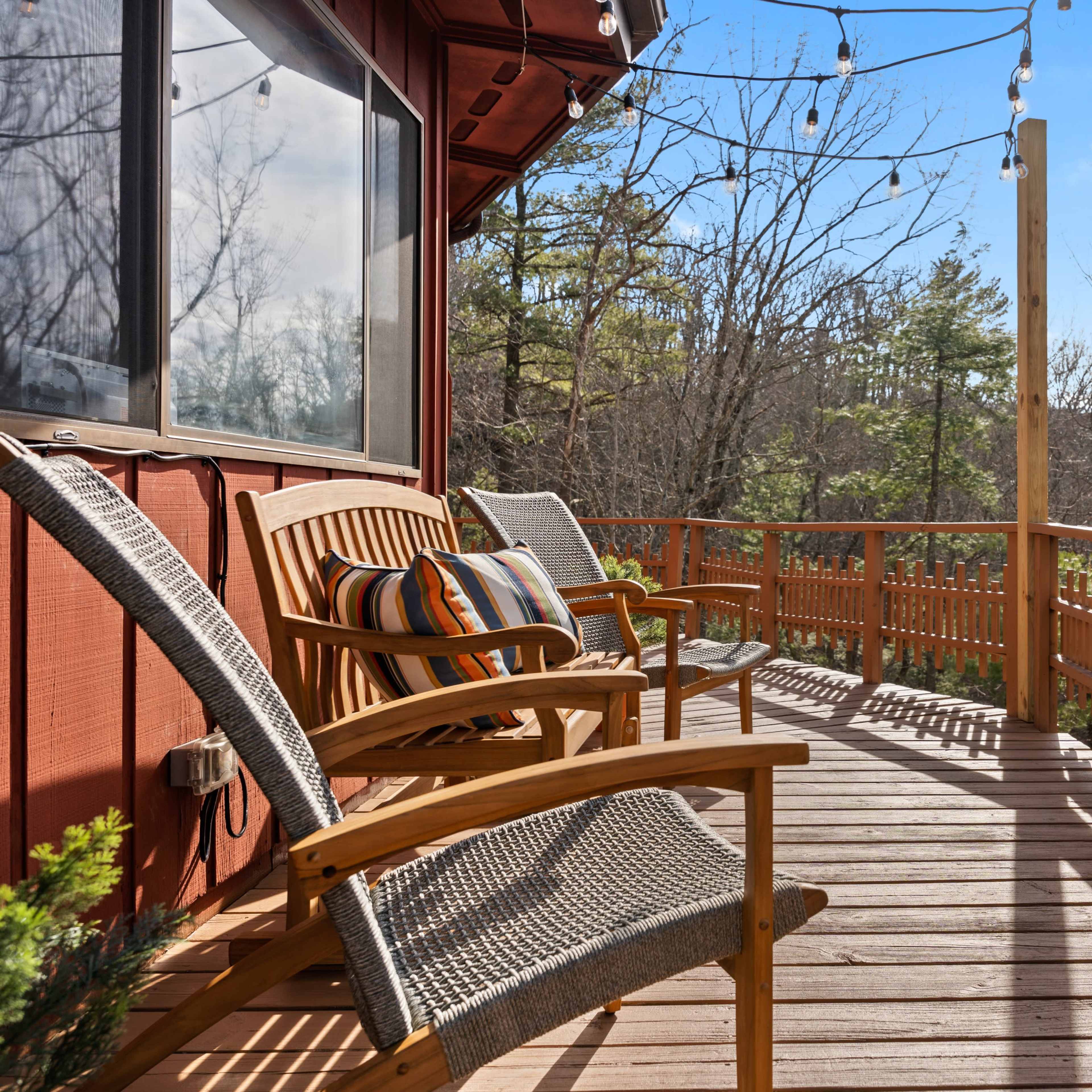 A wooden deck with two chairs and string lights overlooks a tree-lined area.