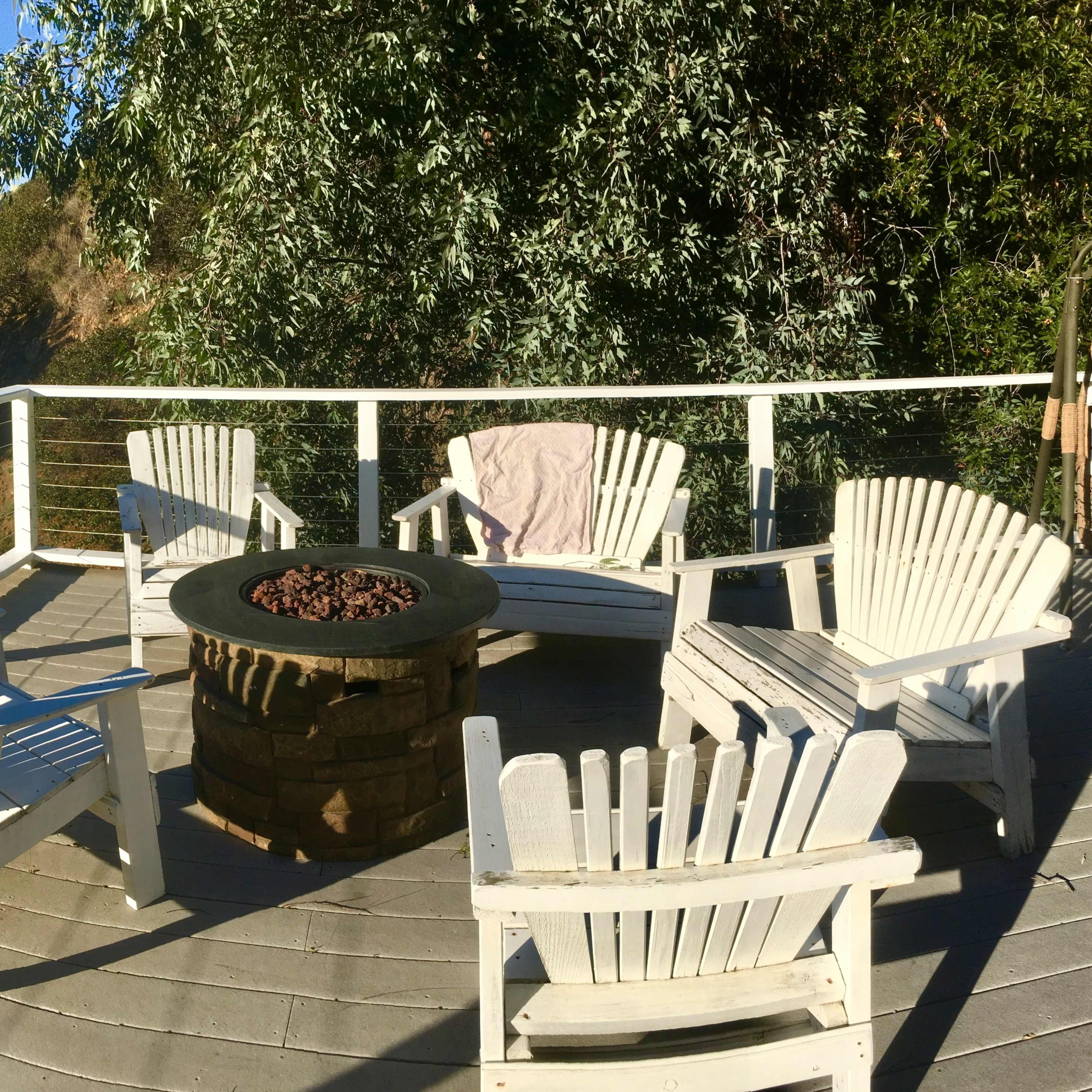A circular fire pit surrounded by six white Adirondack chairs on a wooden deck with a view of green hills in the background.