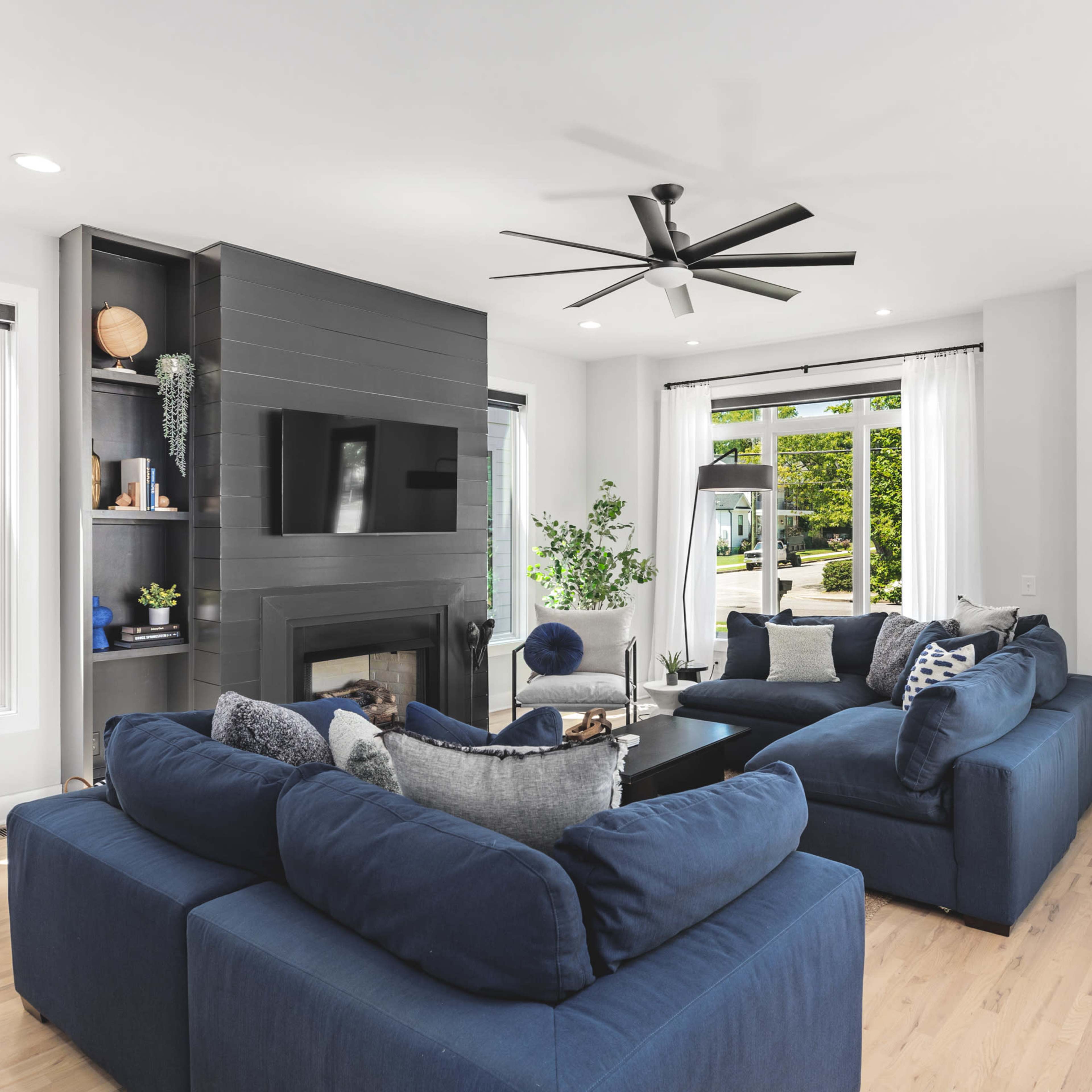 A modern living room features two blue couches arranged around a coffee table, with a fireplace and a large window providing natural light.
