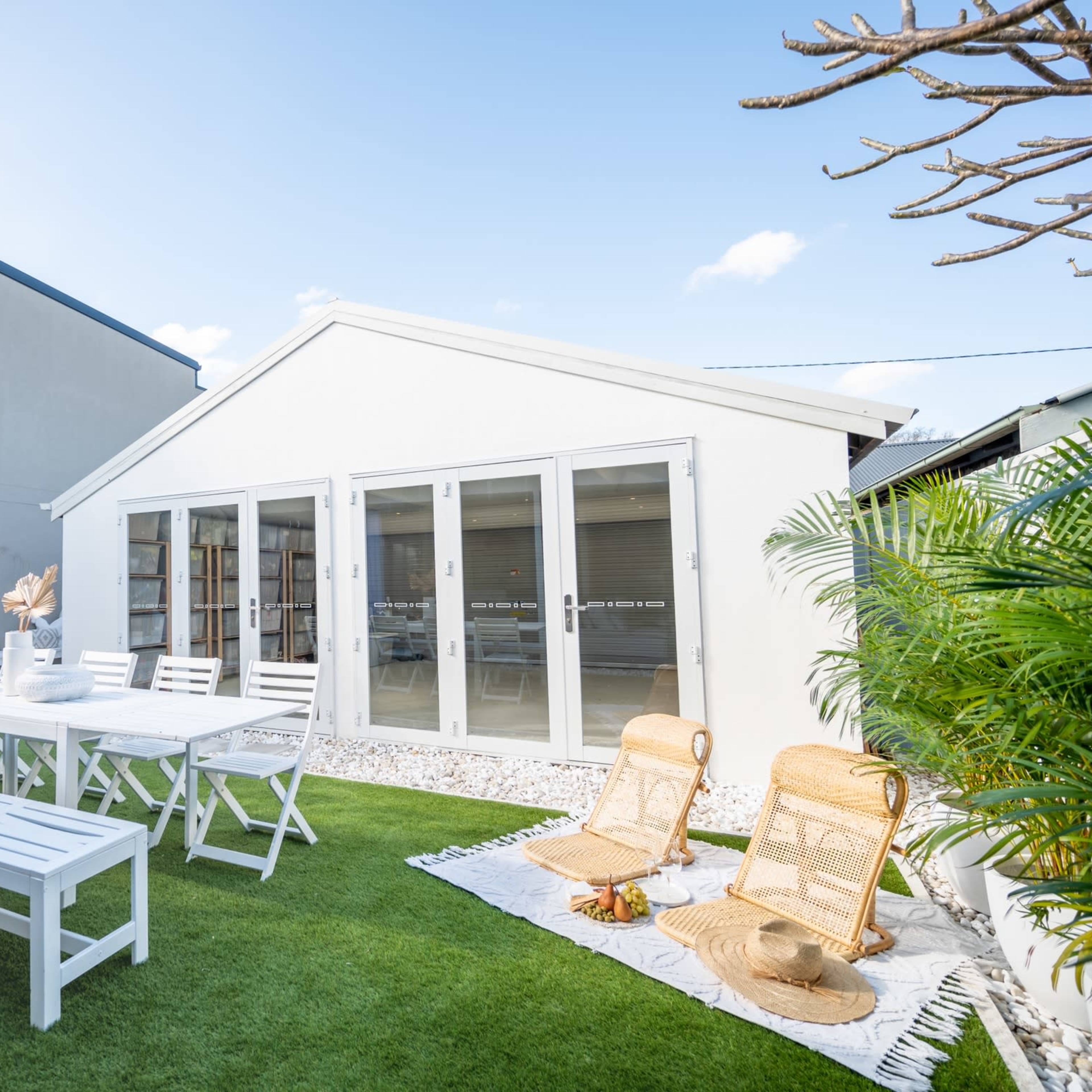 A white garden shed with large glass doors is surrounded by a green lawn, outdoor furniture, and decorative plants.