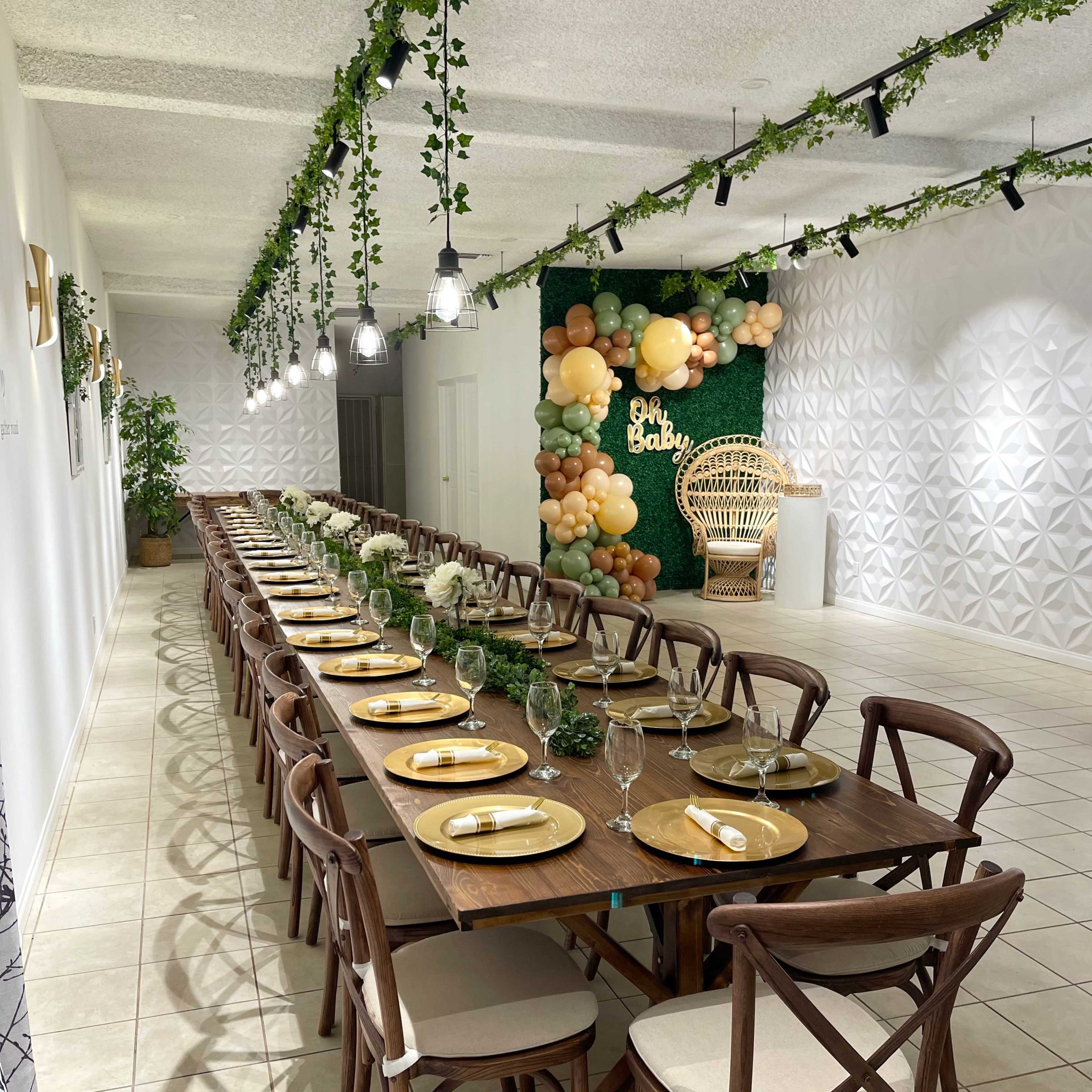 A long, elegantly arranged dining table with wooden chairs set for a special event, adorned with greenery and decorative balloons in a well-lit room.