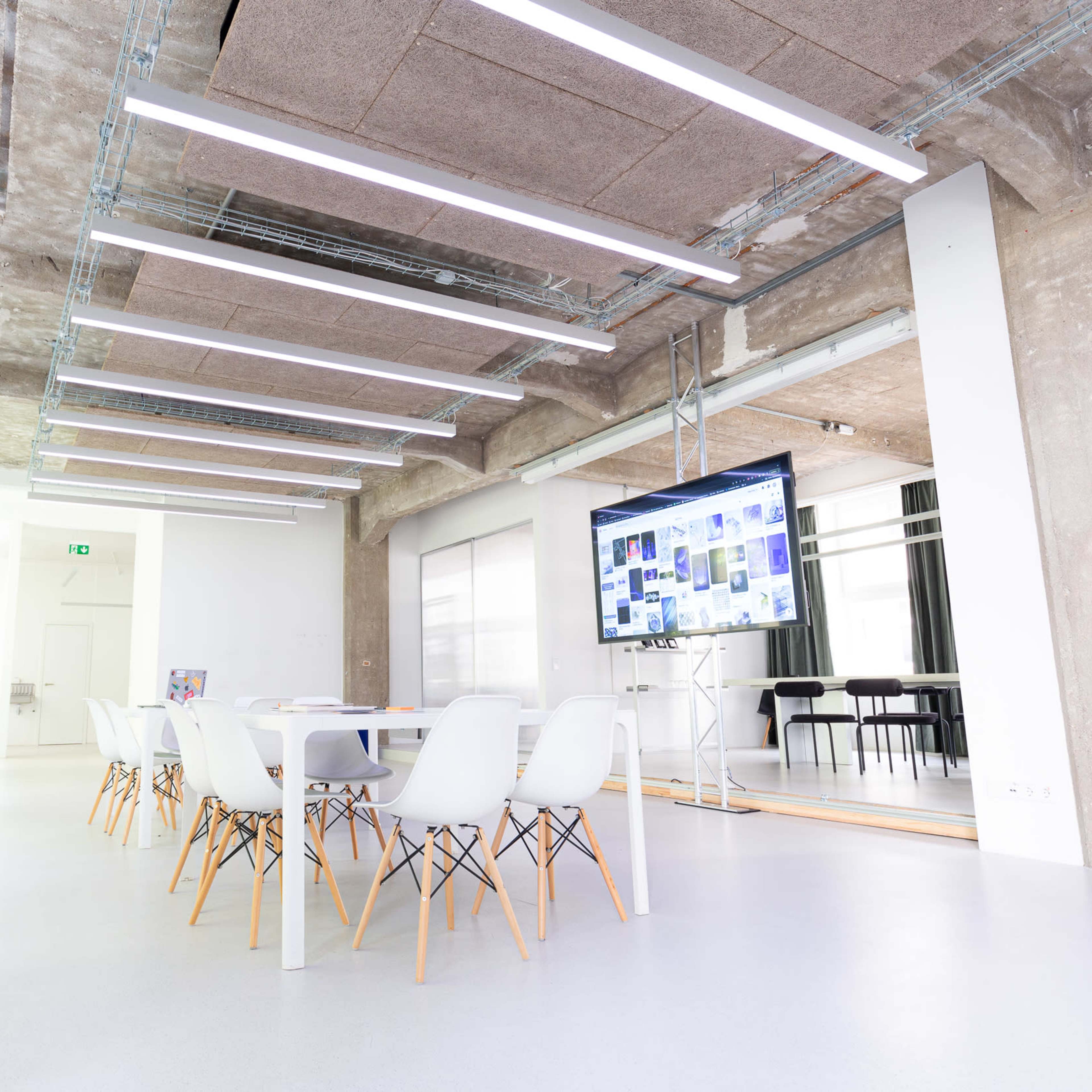 A modern conference room features a long white table surrounded by chairs, with exposed concrete walls and bright overhead lighting.