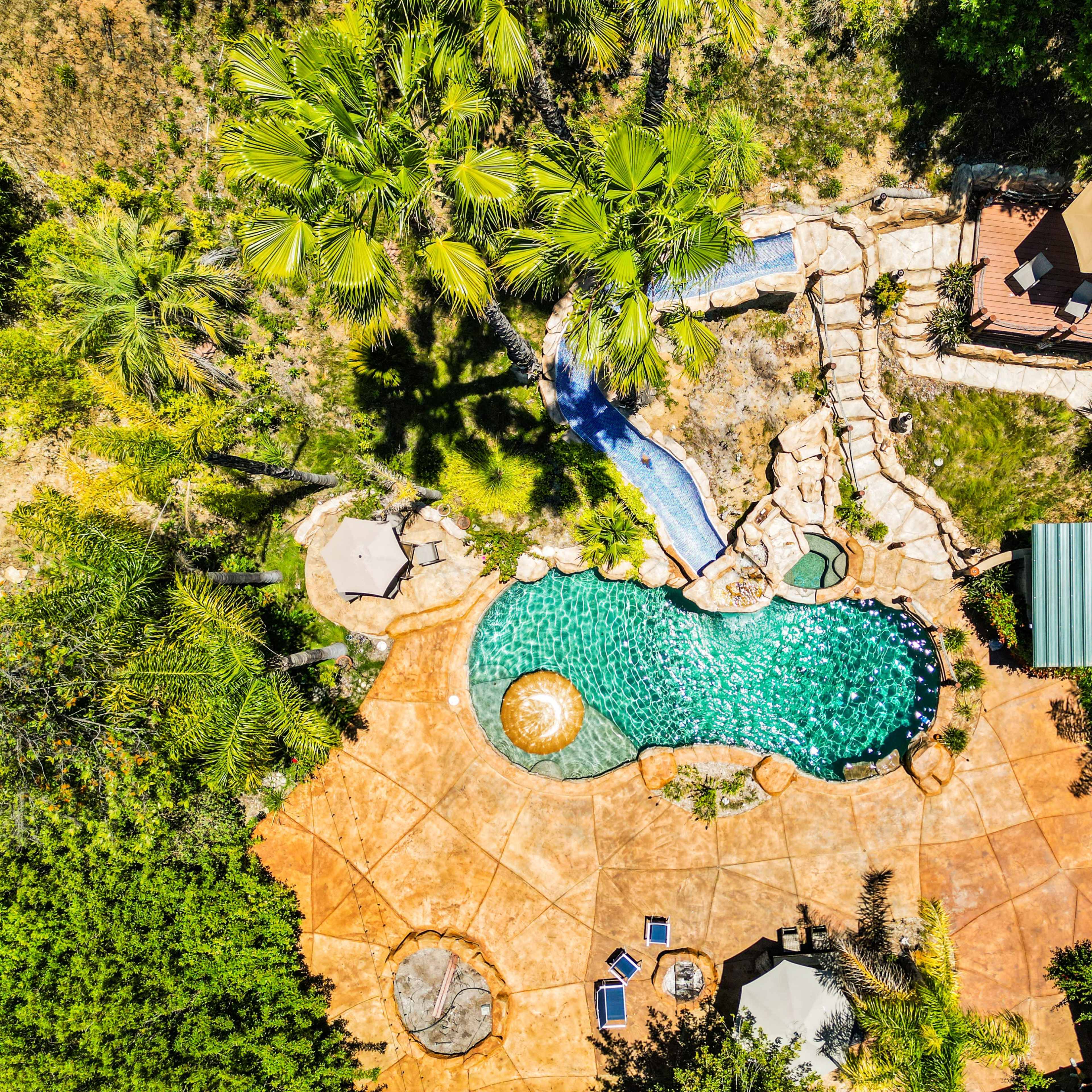 An aerial view of a landscaped backyard featuring a swimming pool surrounded by stone pathways, palm trees, and outdoor seating areas.