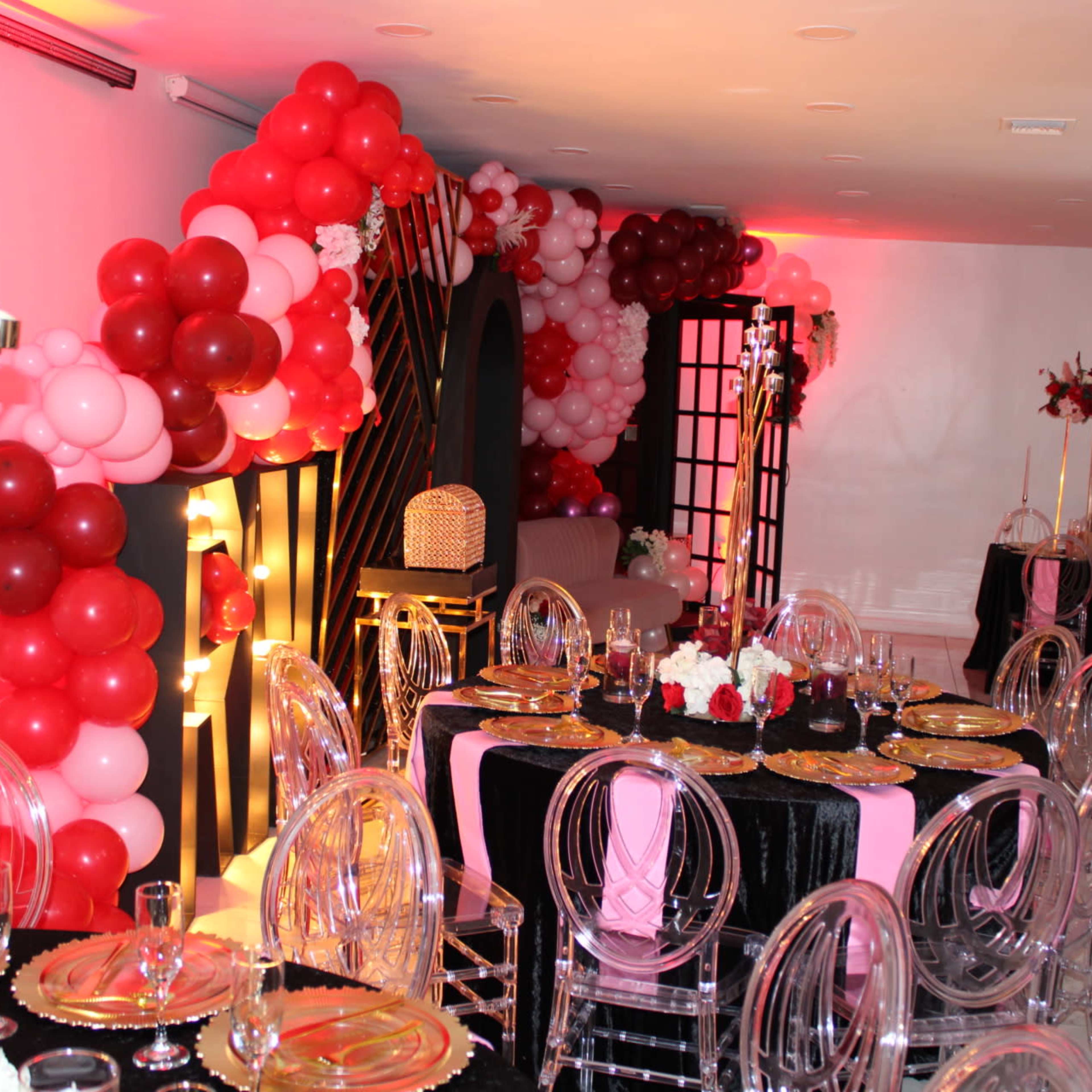 A decorated event space features an arrangement of black and pink tables with transparent chairs, adorned with floral centerpieces and balloon archways in red and pink.