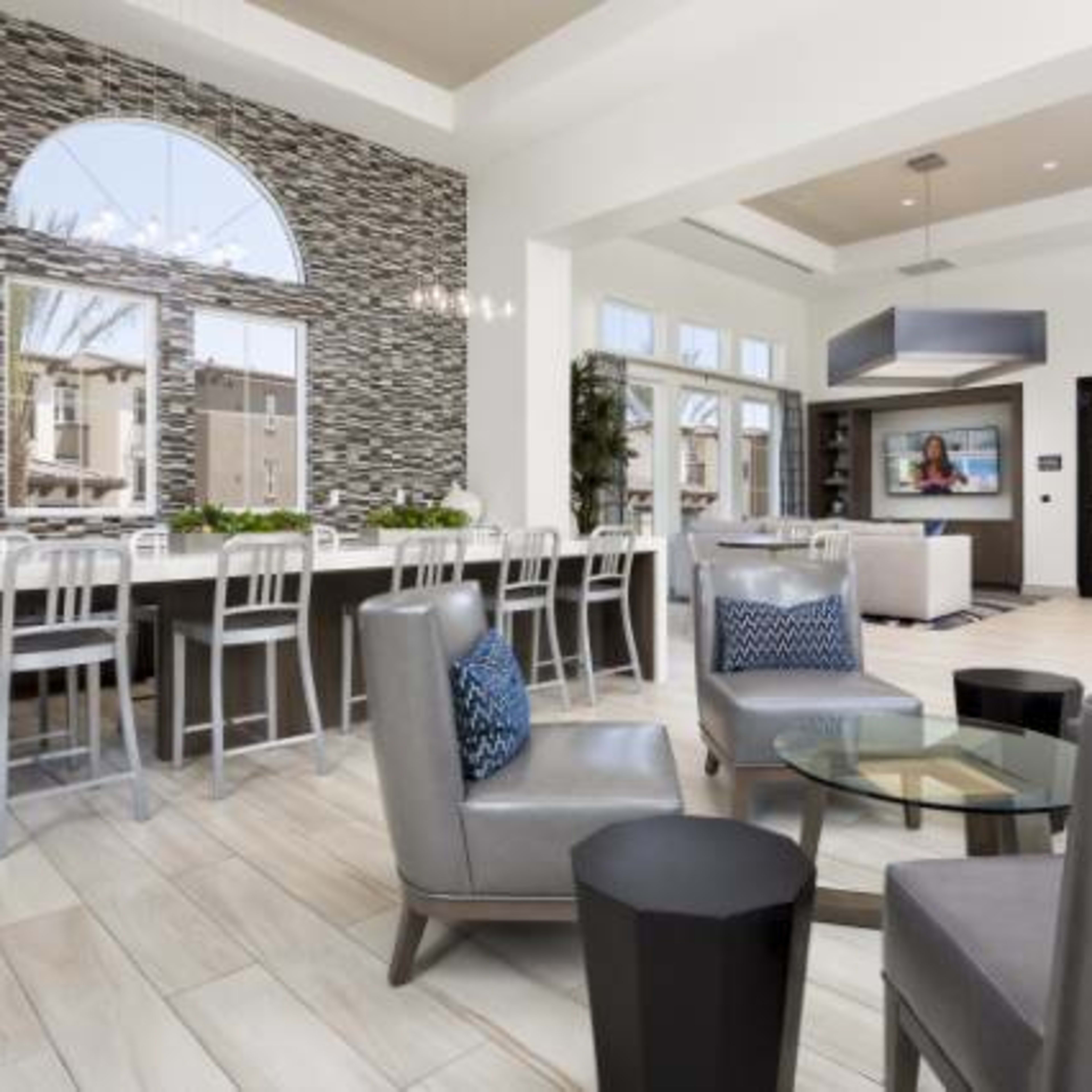 A modern indoor space featuring a kitchen bar with stools, a seating area with gray chairs, and large windows allowing natural light to enter.