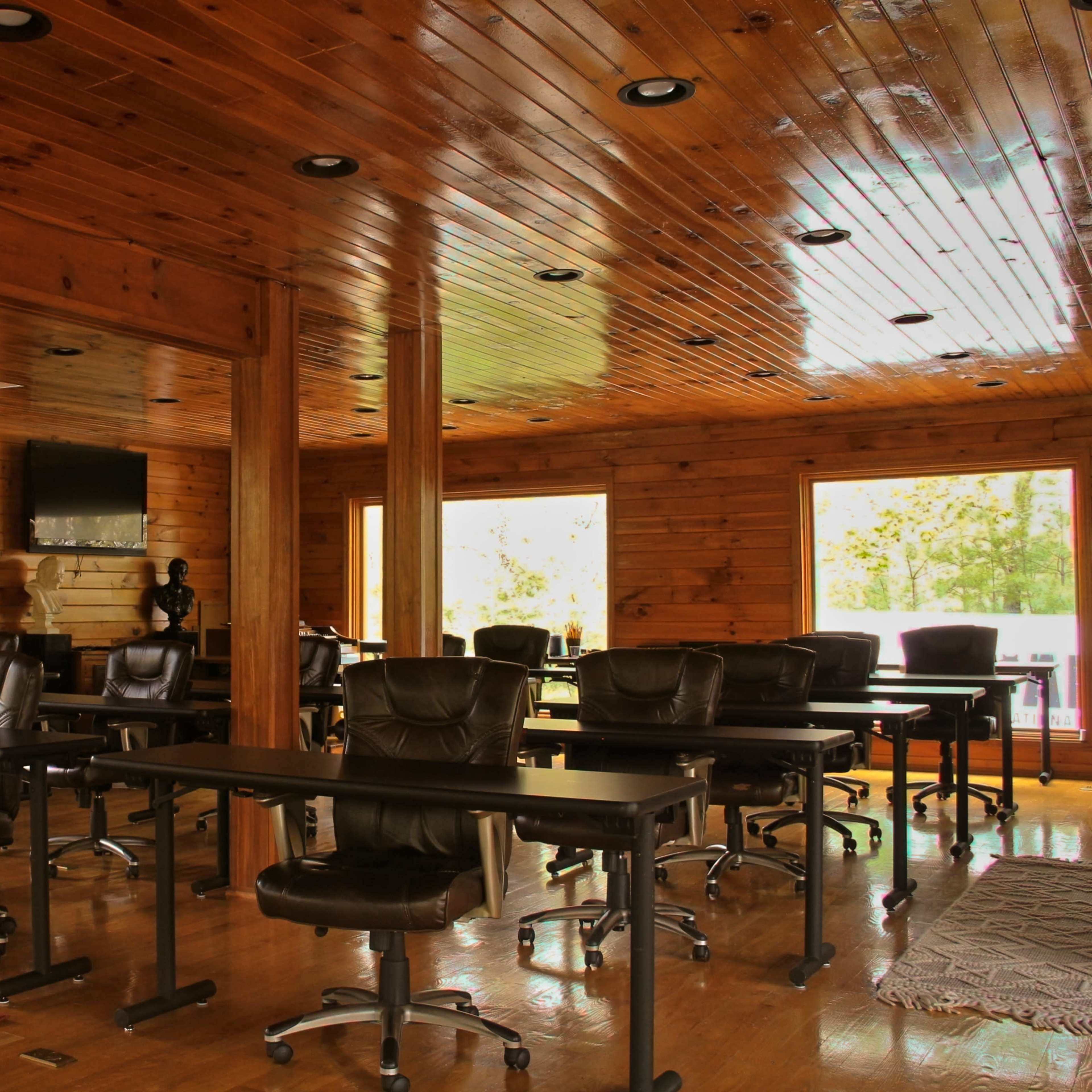 A spacious wooden room set up with several black office chairs and tables, featuring large windows that let in natural light.