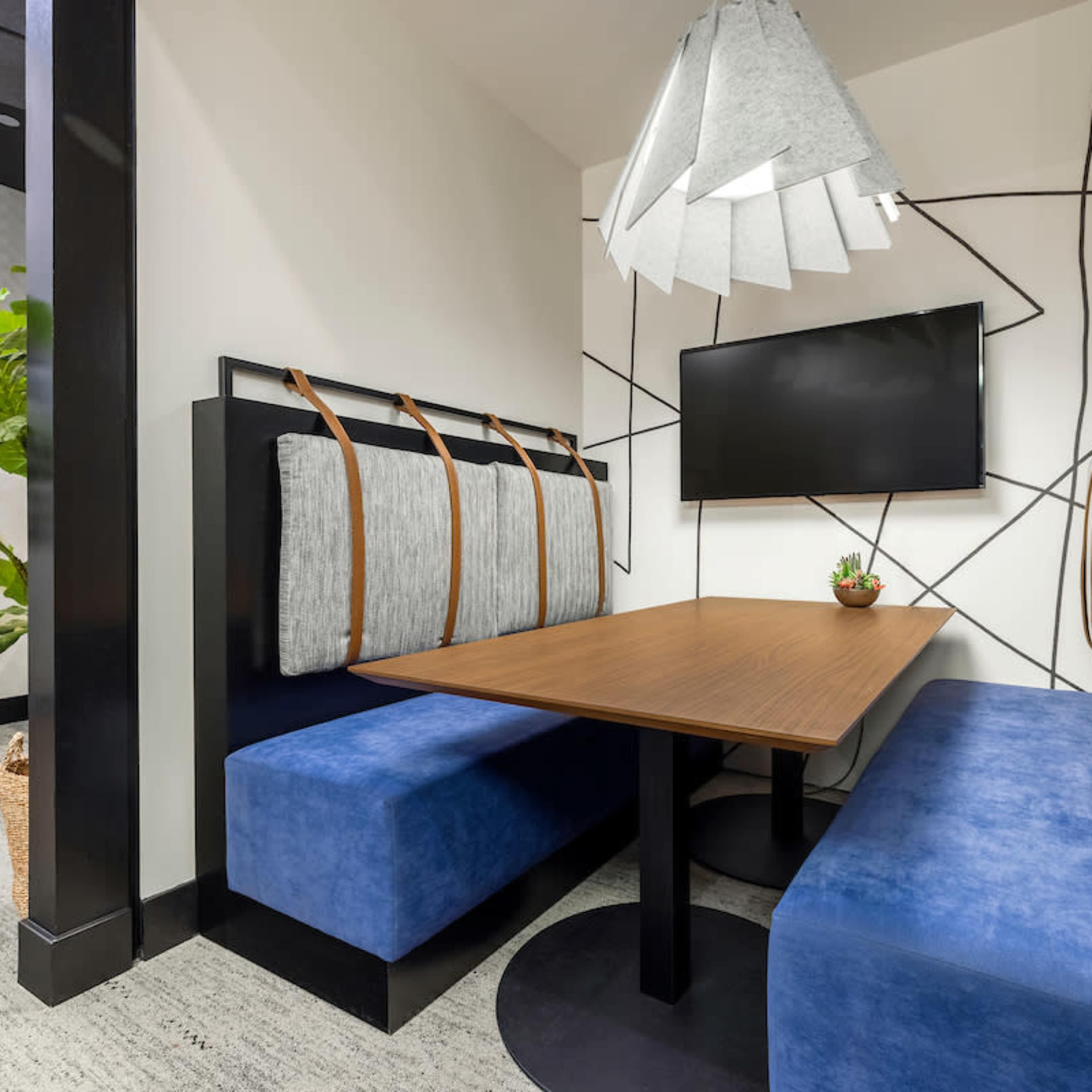 A modern meeting booth with a wooden table, blue upholstered benches, a wall-mounted television, and a decorative plant in a corner.