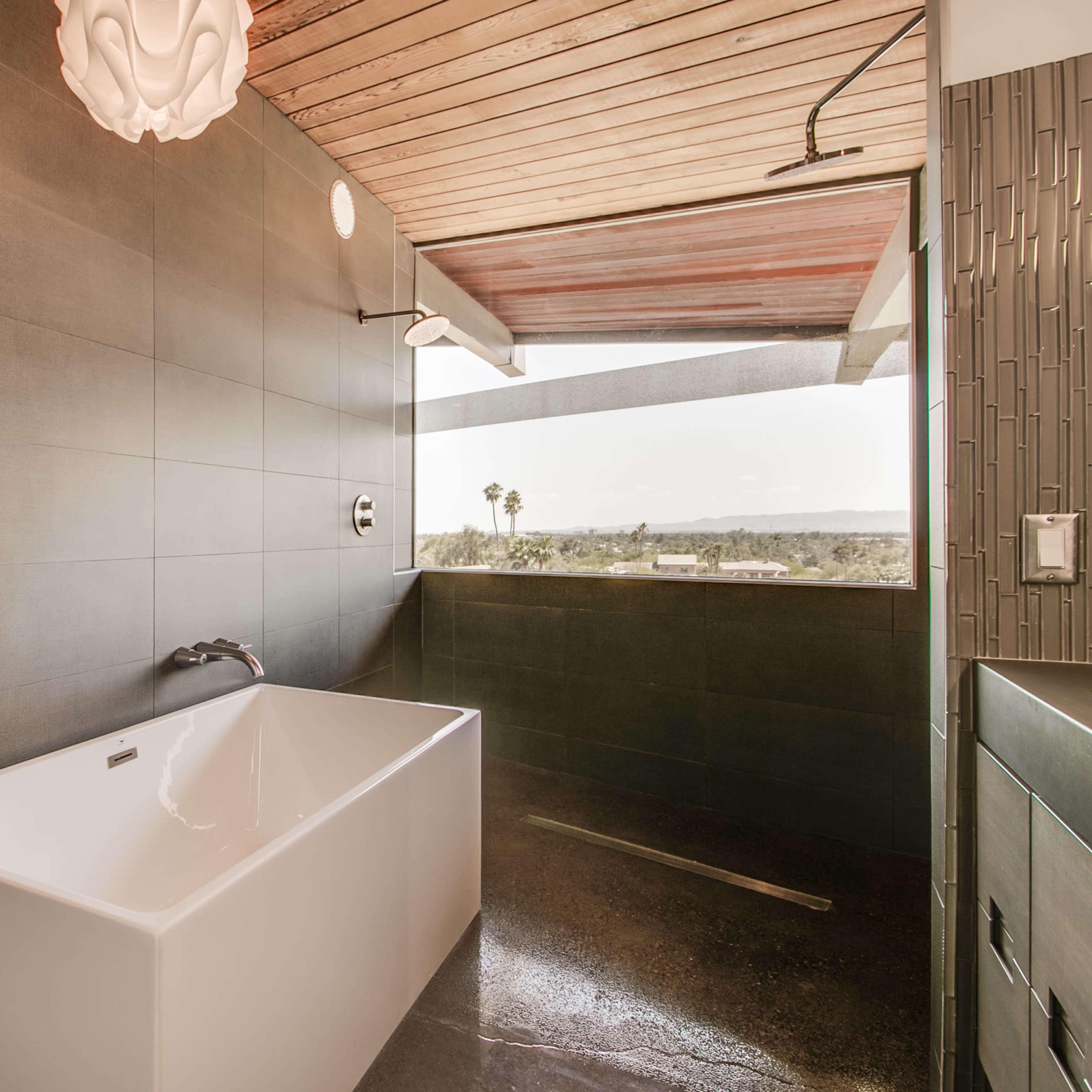 A spacious bathroom features a freestanding white bathtub against a dark tiled wall and large windows with a view.