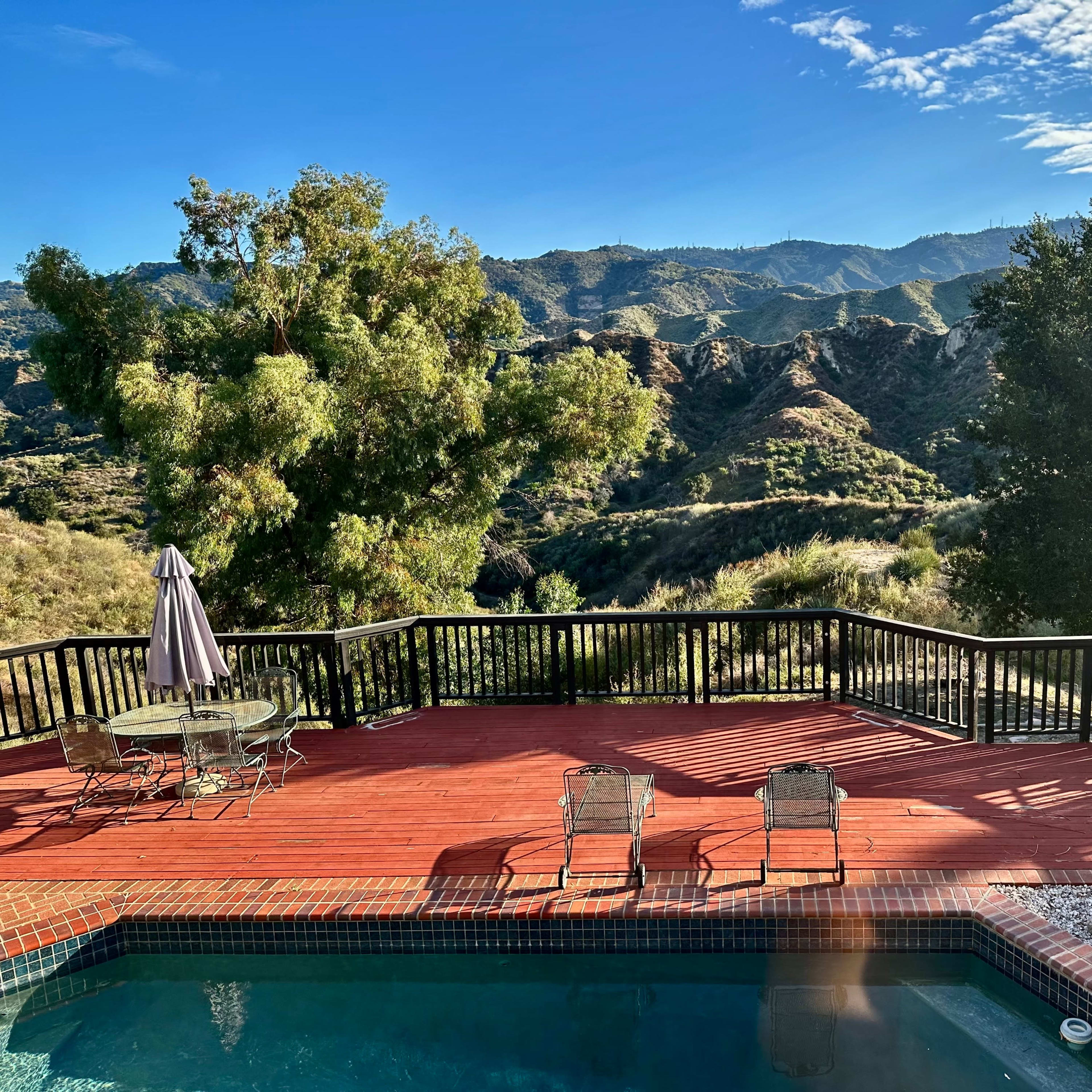 A wooden deck with two chairs and a table overlooks a pool and a mountainous landscape under a blue sky with scattered clouds.