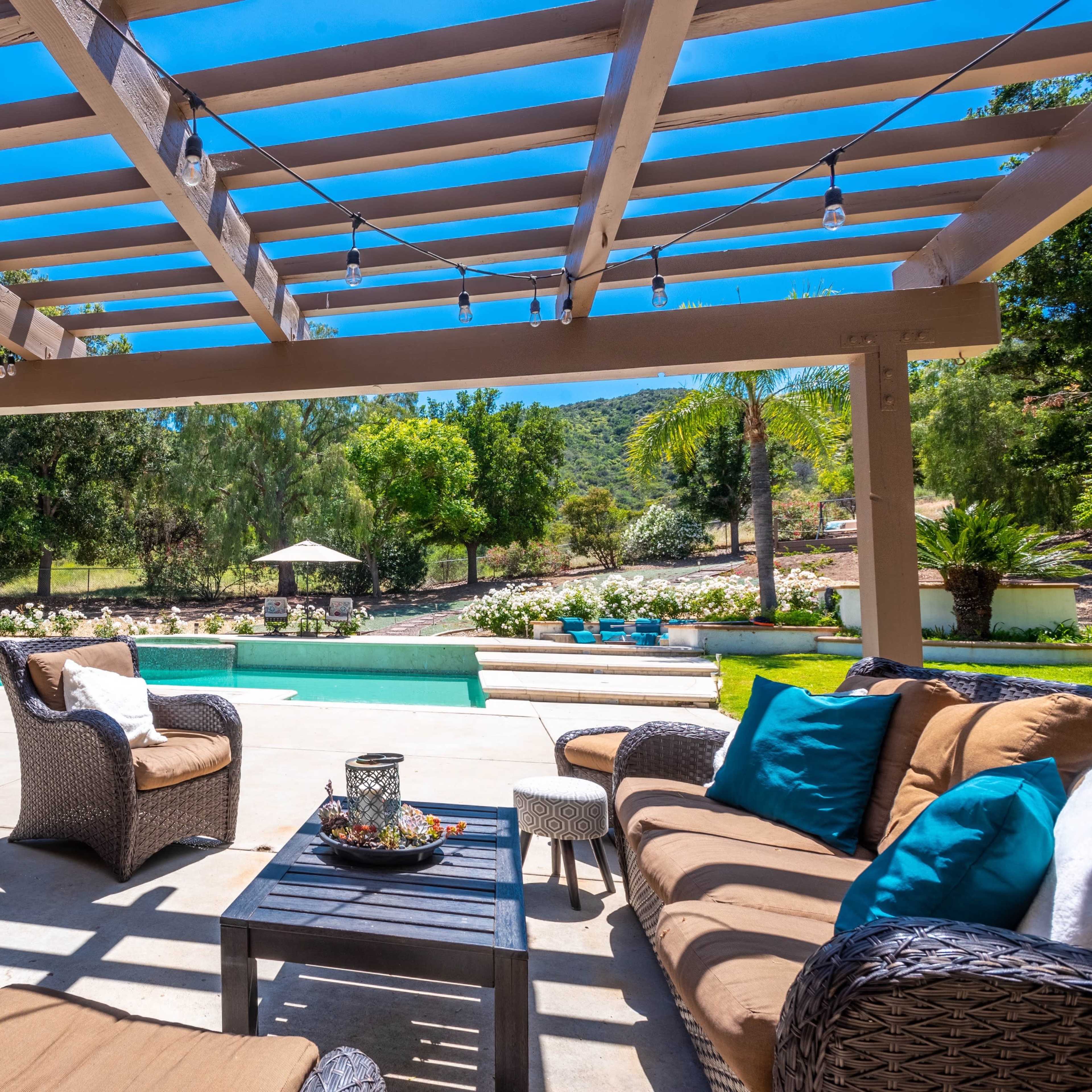 A patio area features wicker furniture arranged around a table, overlooking a pool surrounded by greenery and decorative plants.