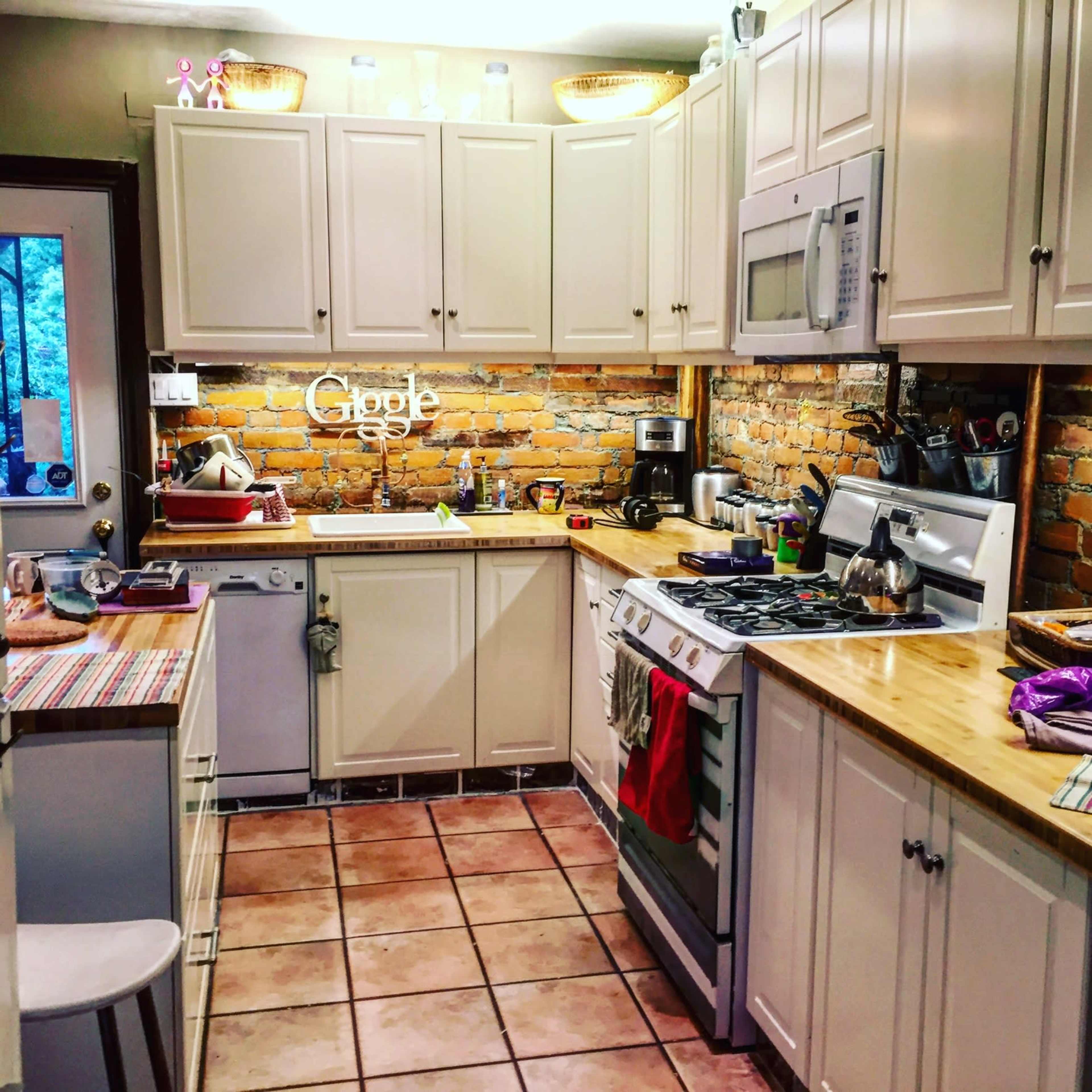 A compact kitchen features wooden countertops, white cabinets, and exposed brick walls, with appliances neatly arranged and several kitchen utensils displayed.