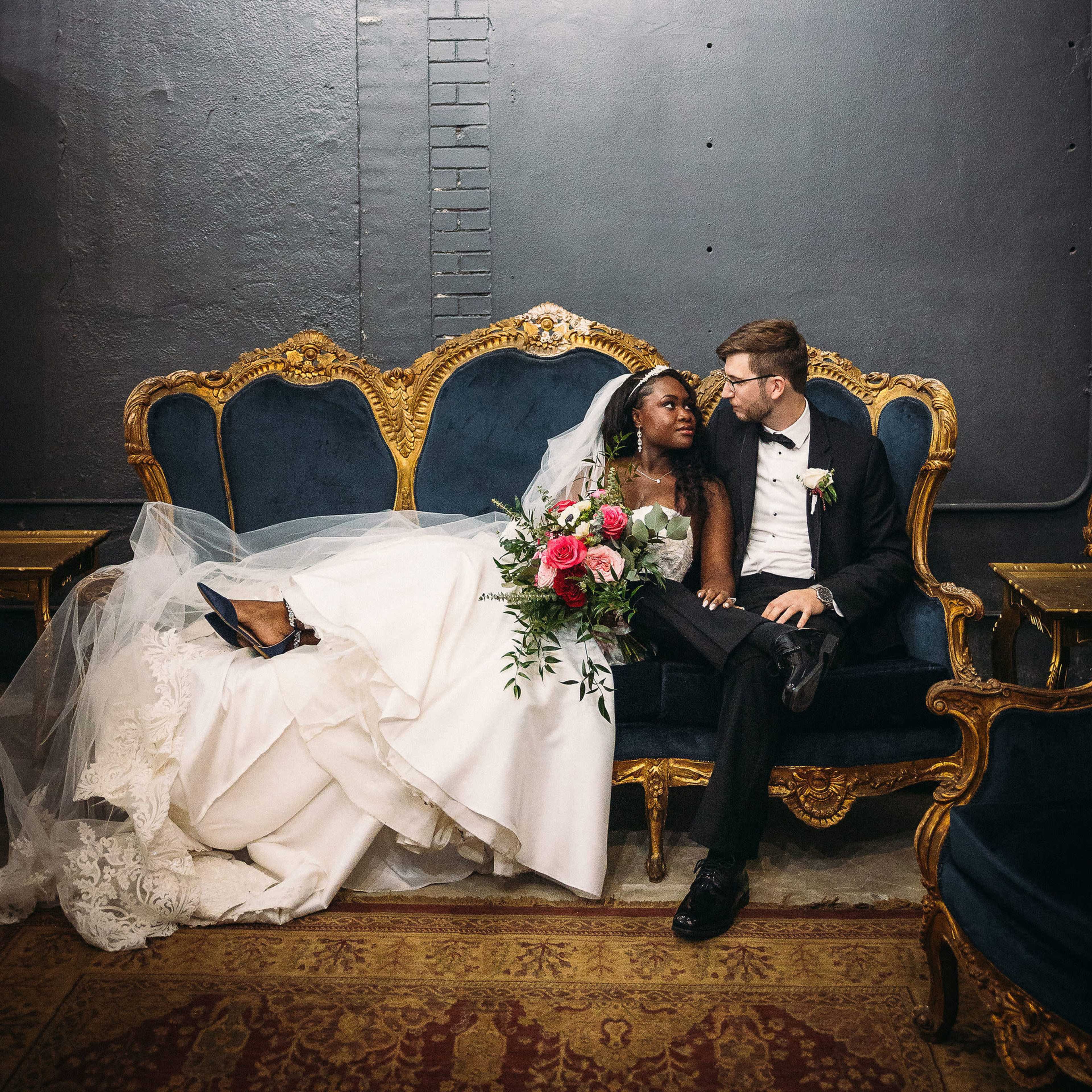 A bride and groom sit closely together on a vintage blue velvet couch, surrounded by dim lighting and dark walls.