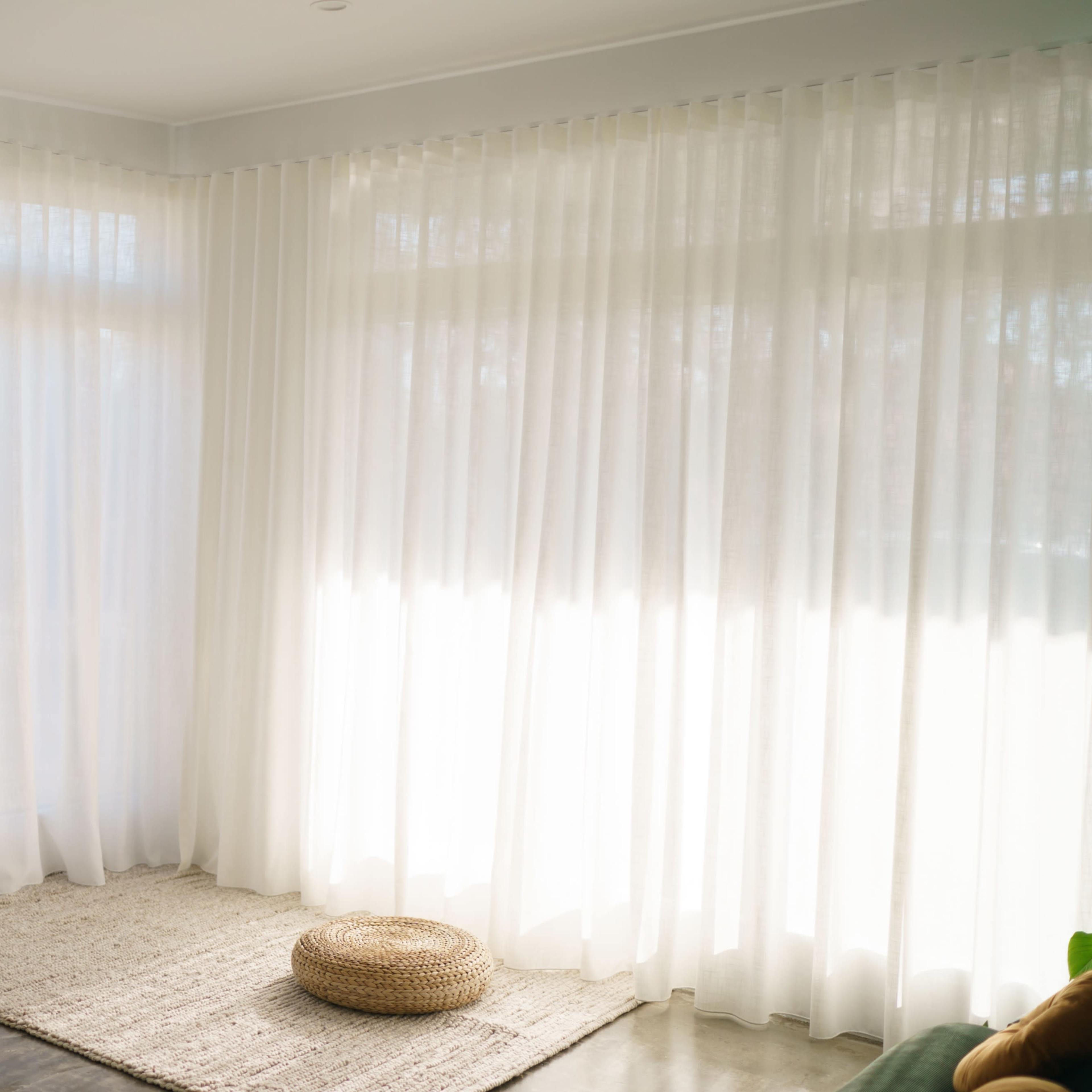 A bright room features sheer white curtains, a woven rug, and a circular pouf resting on a polished concrete floor.