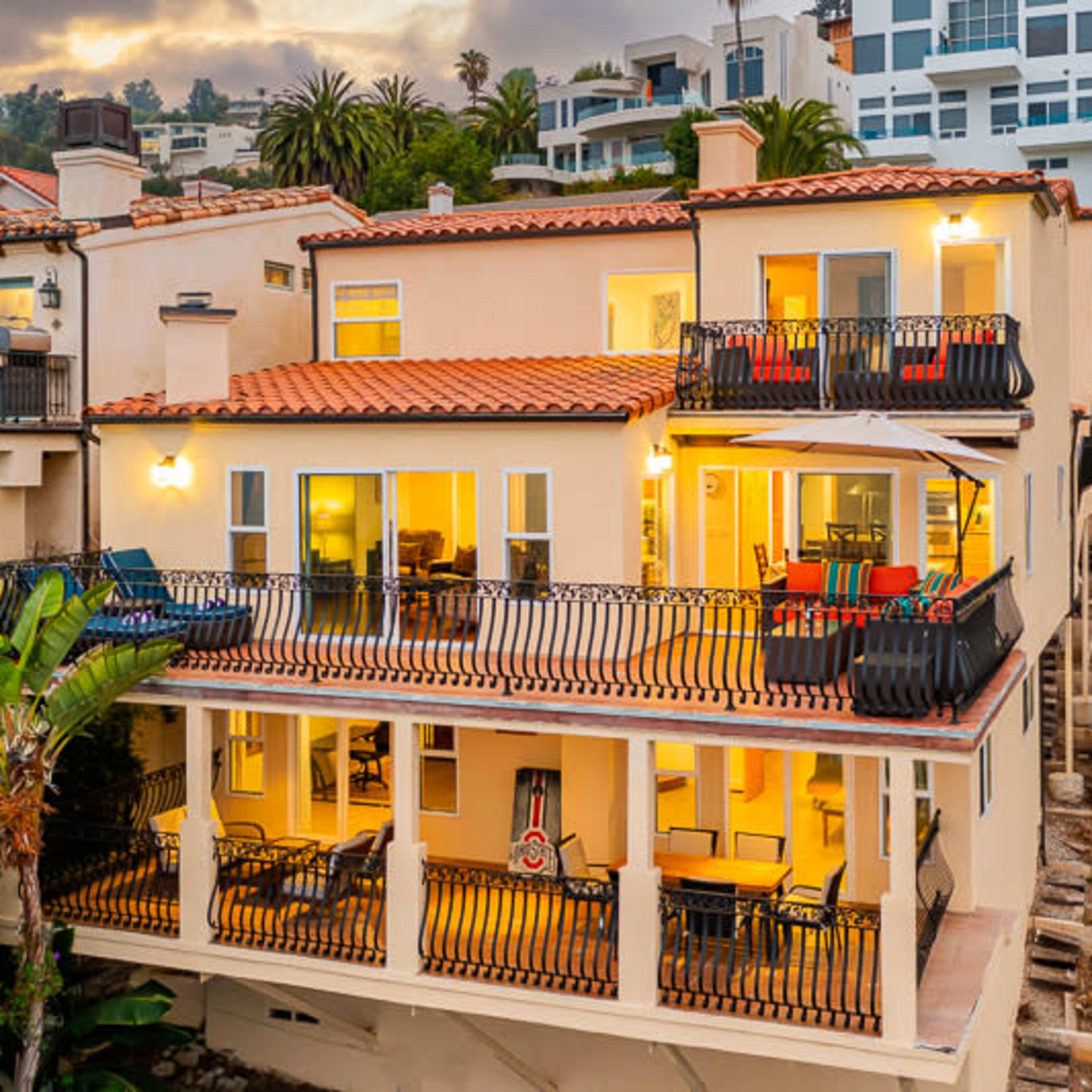 A multi-level beachfront house with a terracotta-tile roof, large balconies, and palm trees surrounding the property.