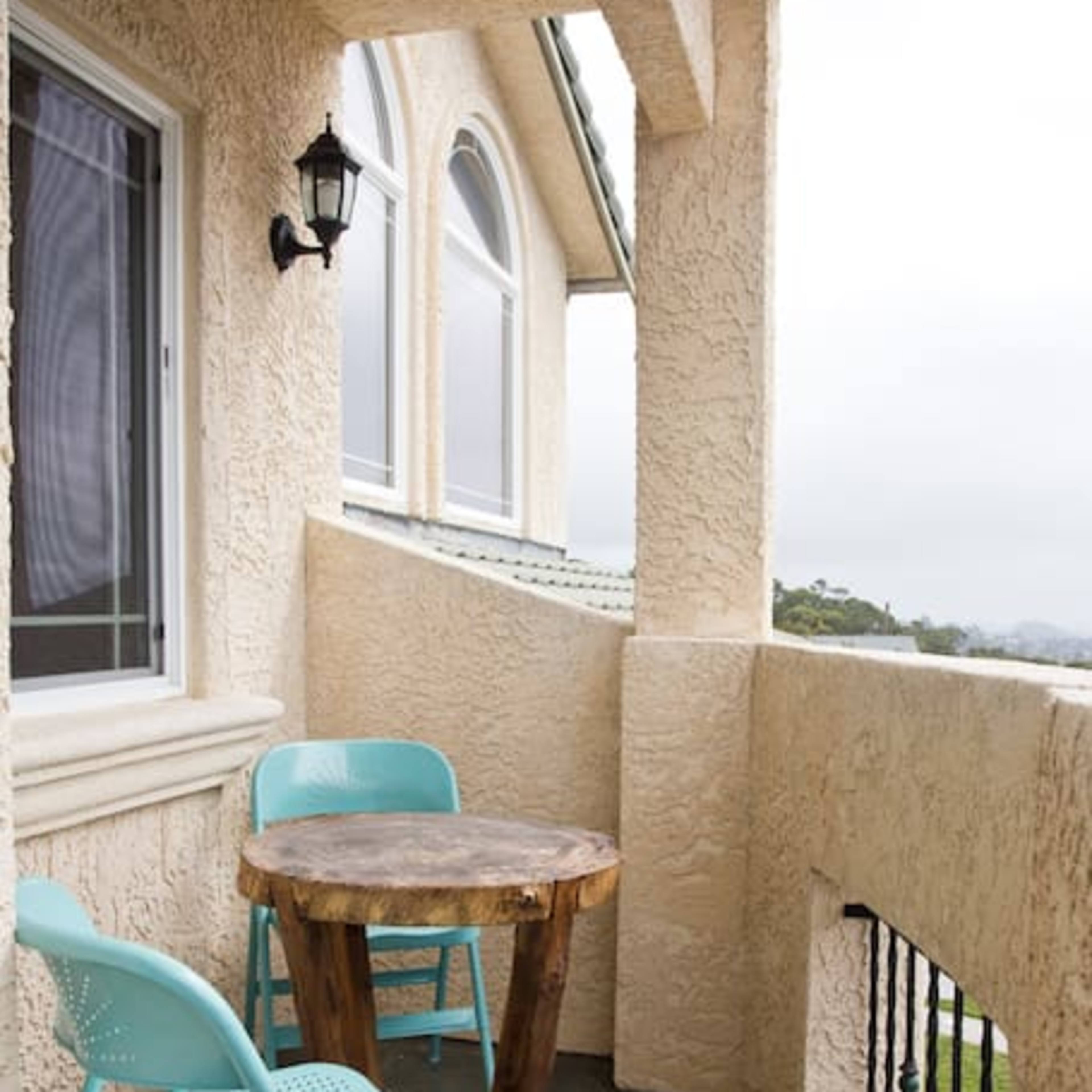 A small balcony with a round wooden table and two turquoise chairs, featuring a stone floor and a wall-mounted light fixture.