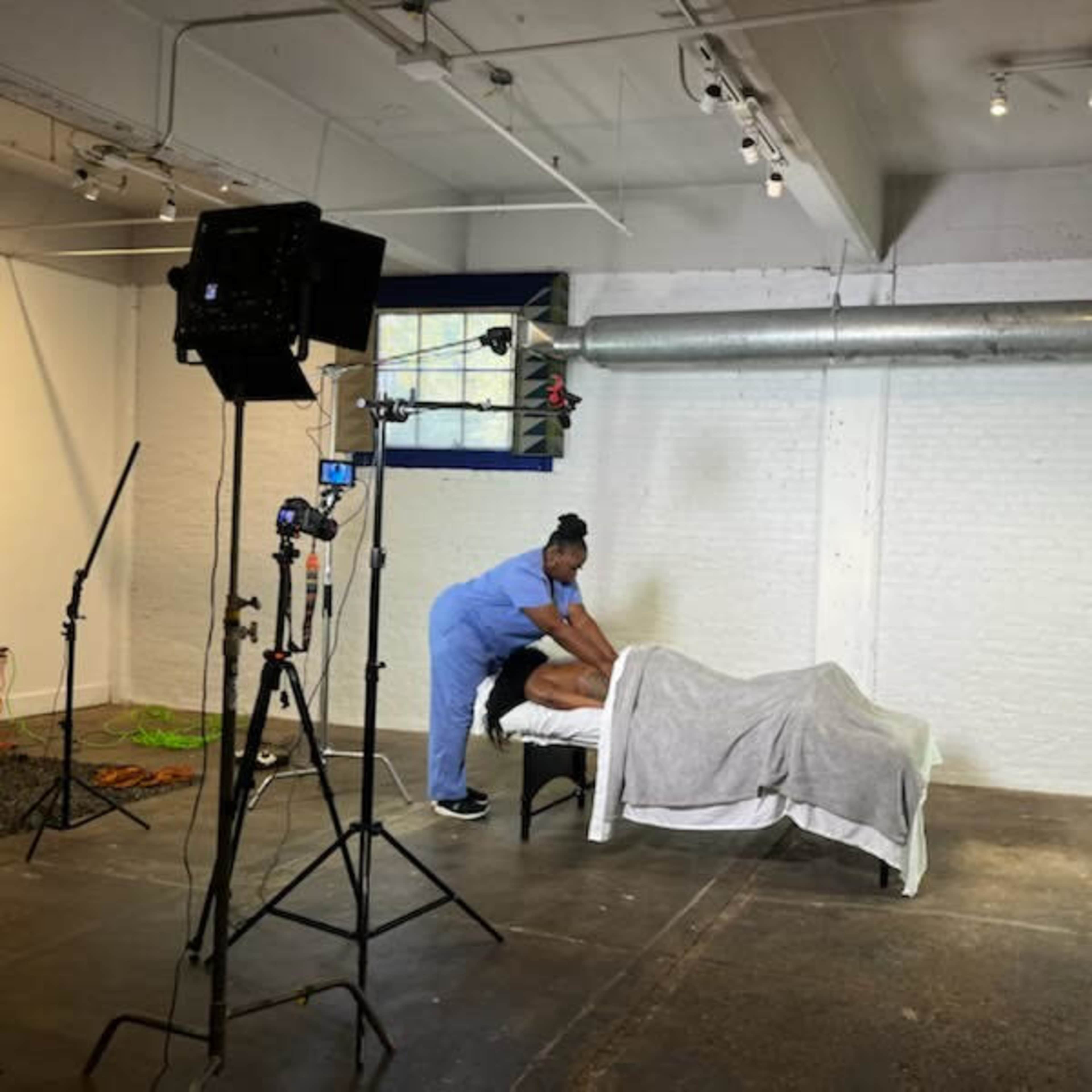 A person in blue scrubs is giving a massage to an individual covered by a gray blanket on a treatment table in a well-lit room.