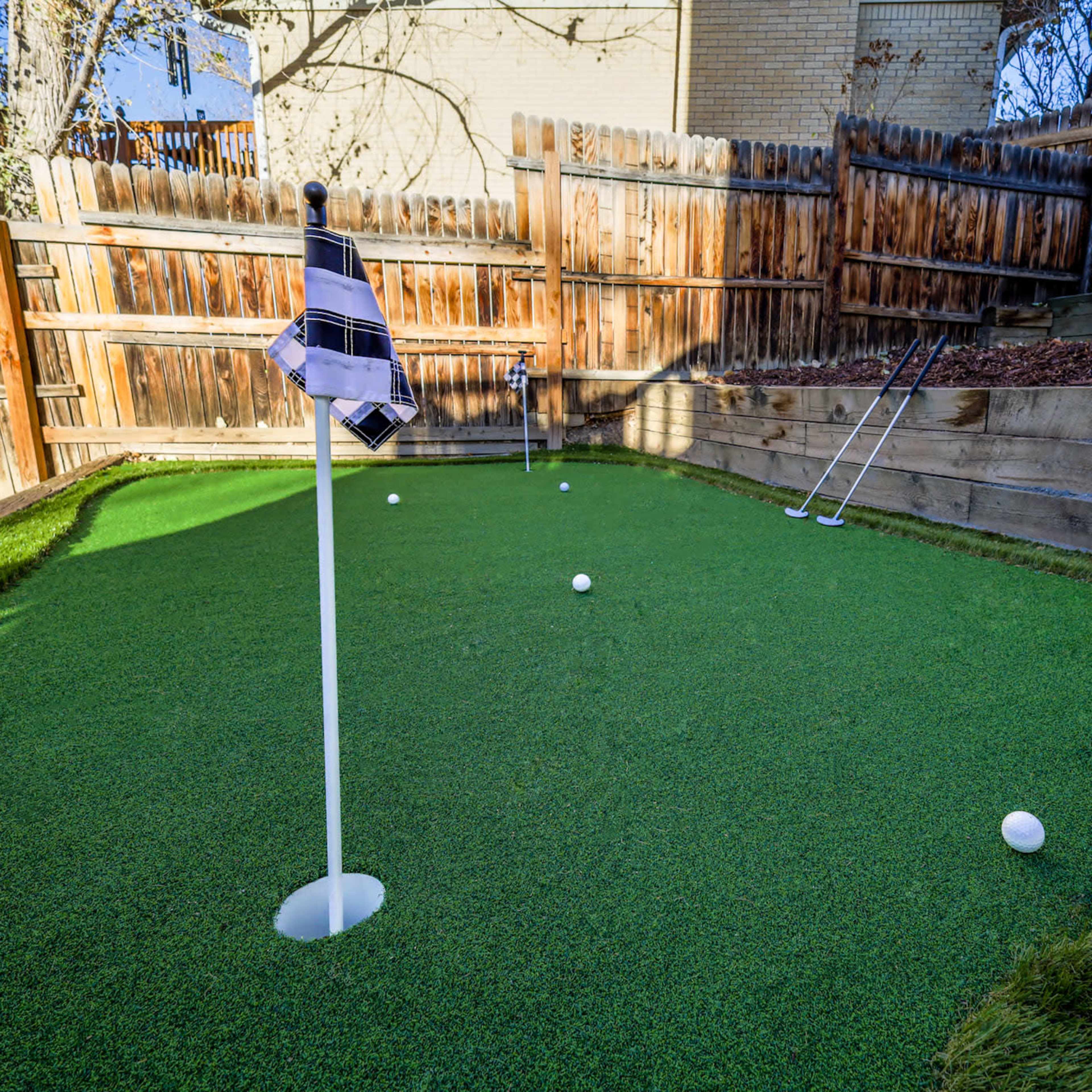 A small putting green in a backyard, featuring a flag in a hole and several golf balls on the artificial grass surface.