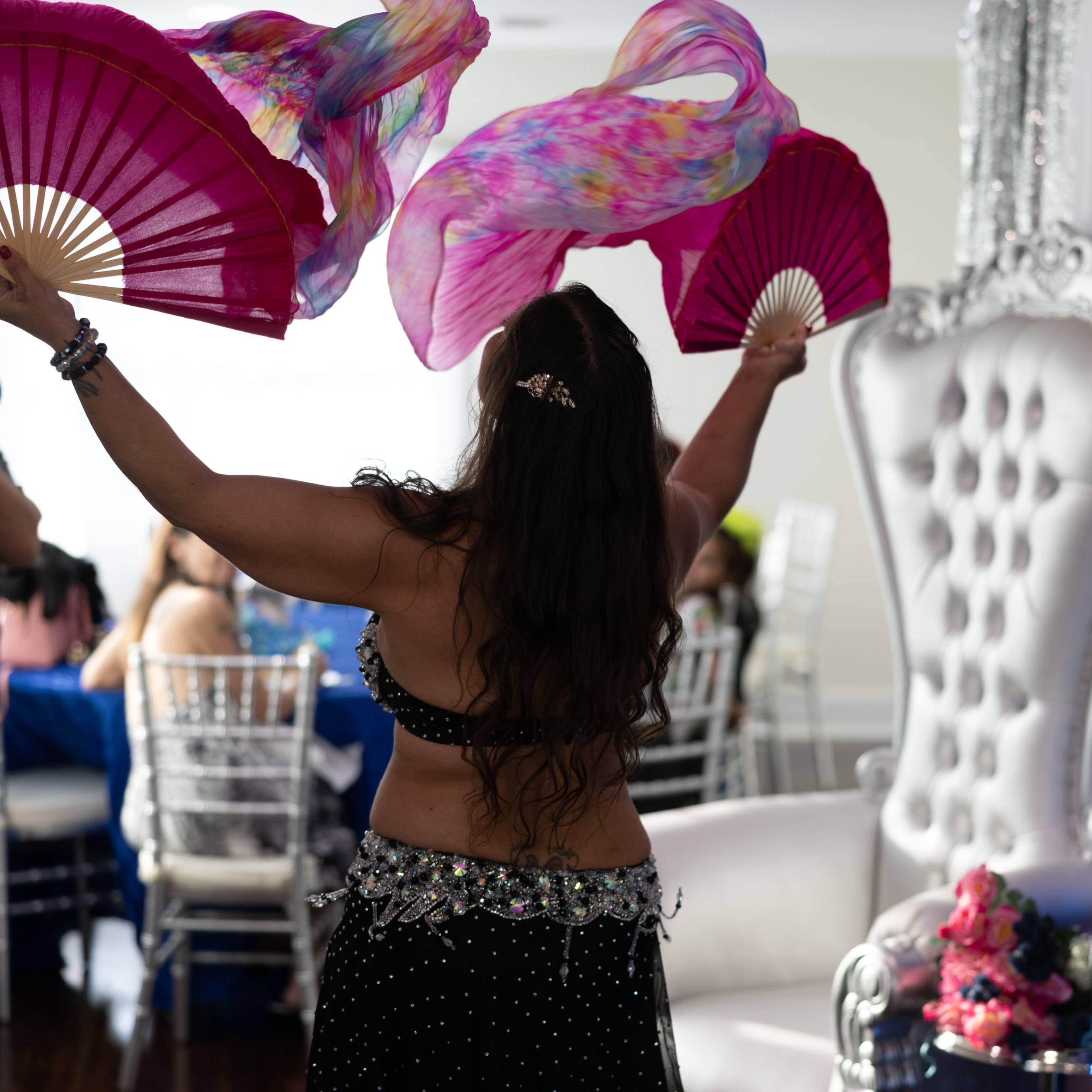 A dancer performing with colorful fans in front of a decorated backdrop featuring a white throne and blue balloons.