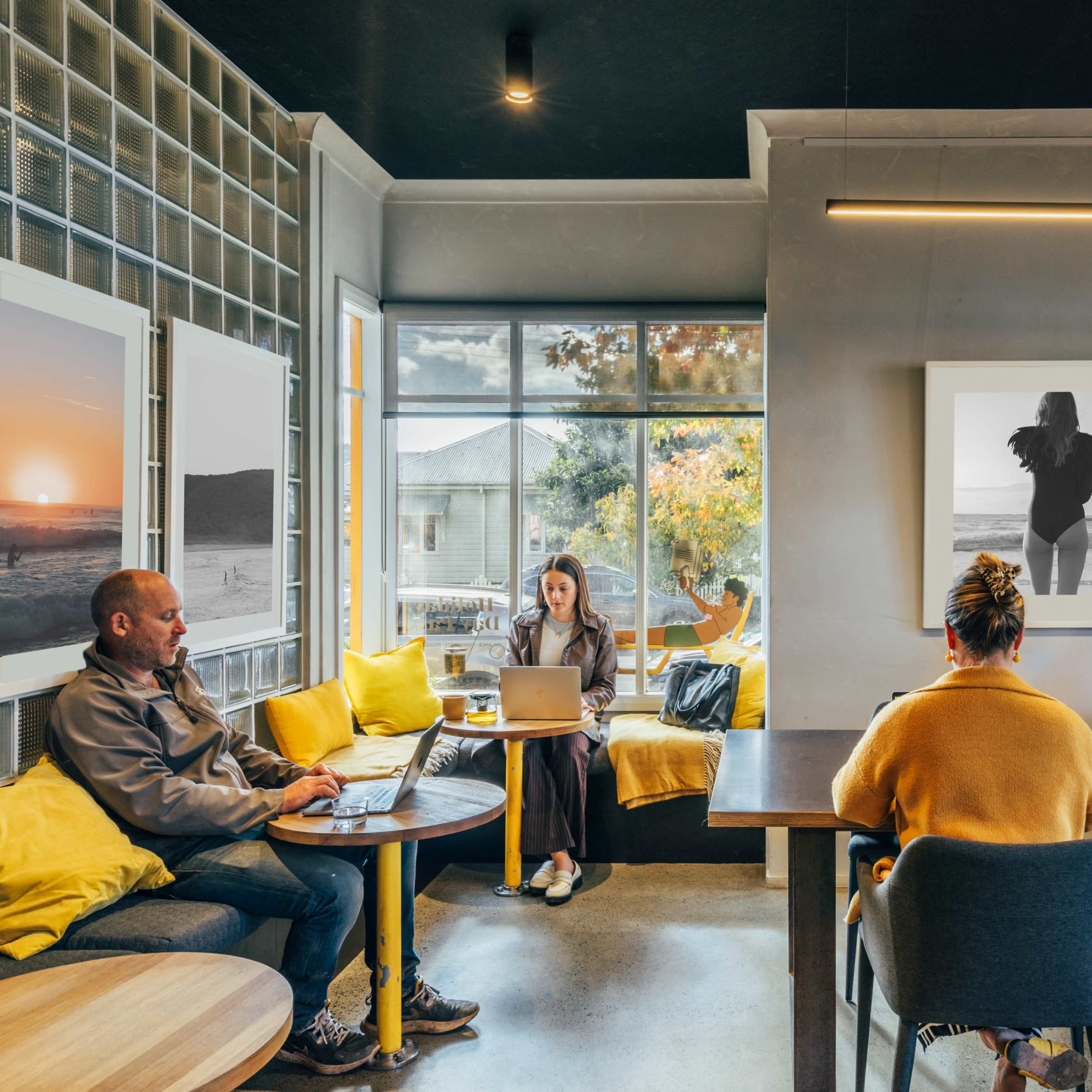 A cozy café interior features several people working on laptops at different tables, surrounded by large windows and artistic wall decor.