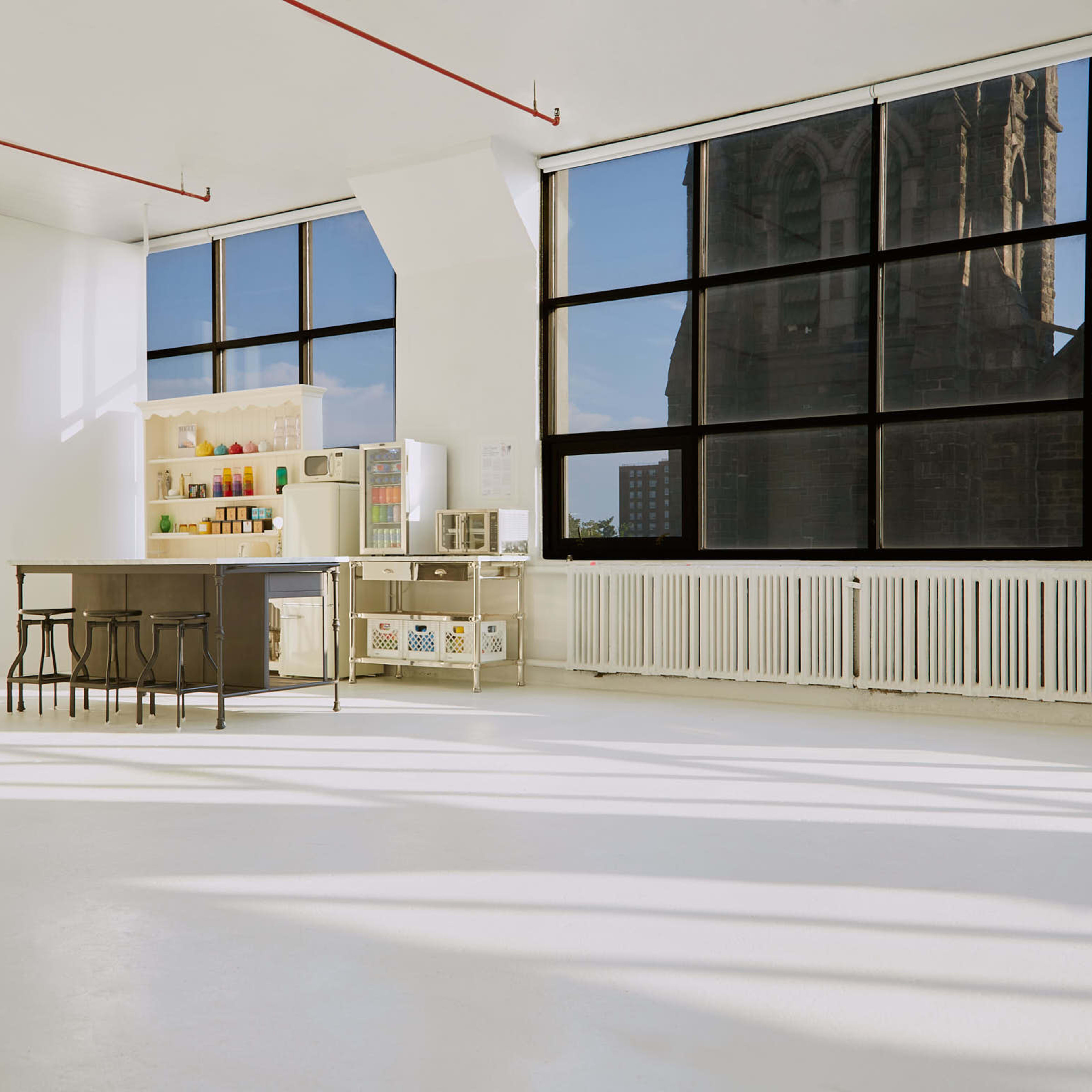 A bright, empty room features a kitchen island and a shelf of colorful jars, with large windows overlooking a stone church.