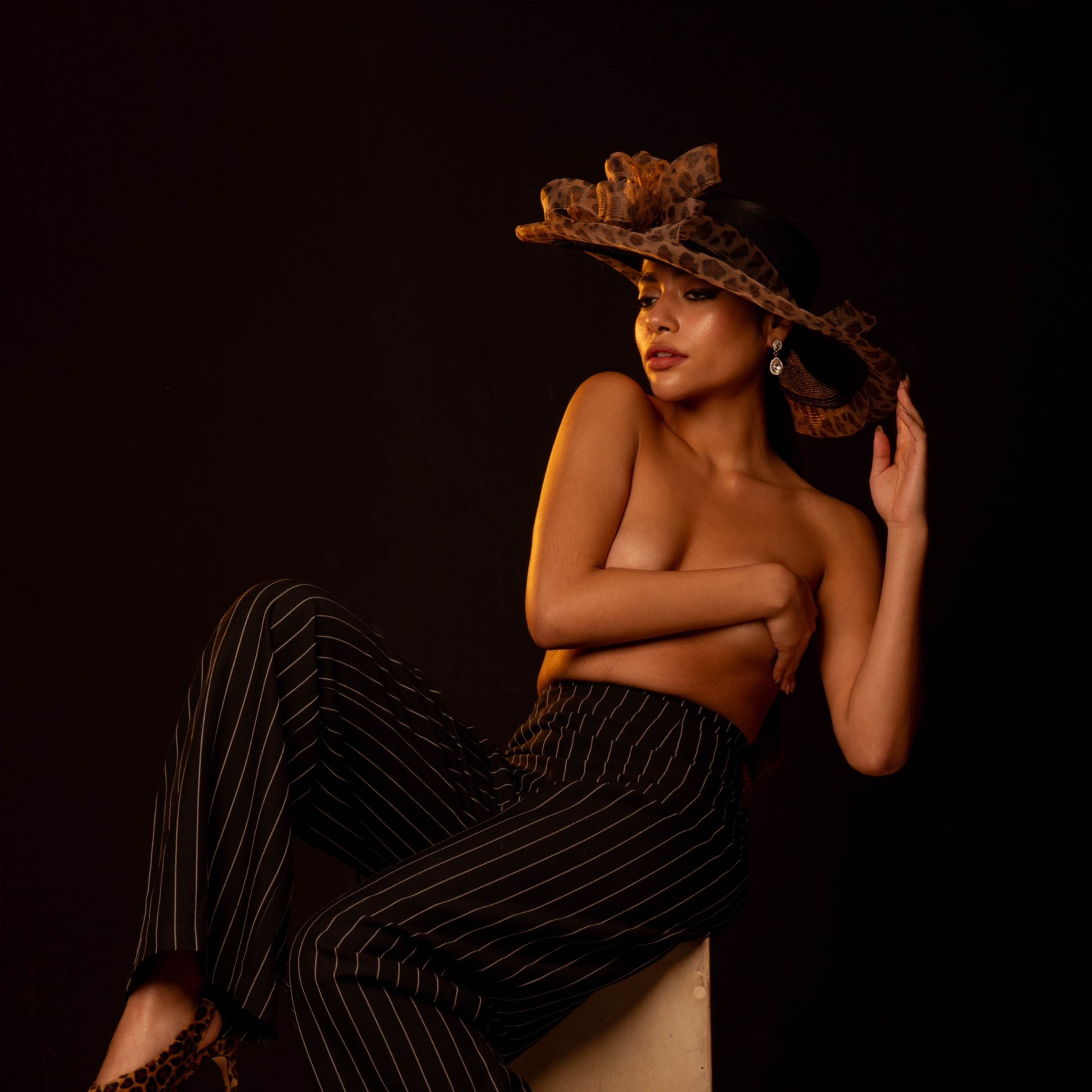 A model poses on a wooden block wearing pinstriped pants, a large hat, and leopard print shoes, against a dark background.