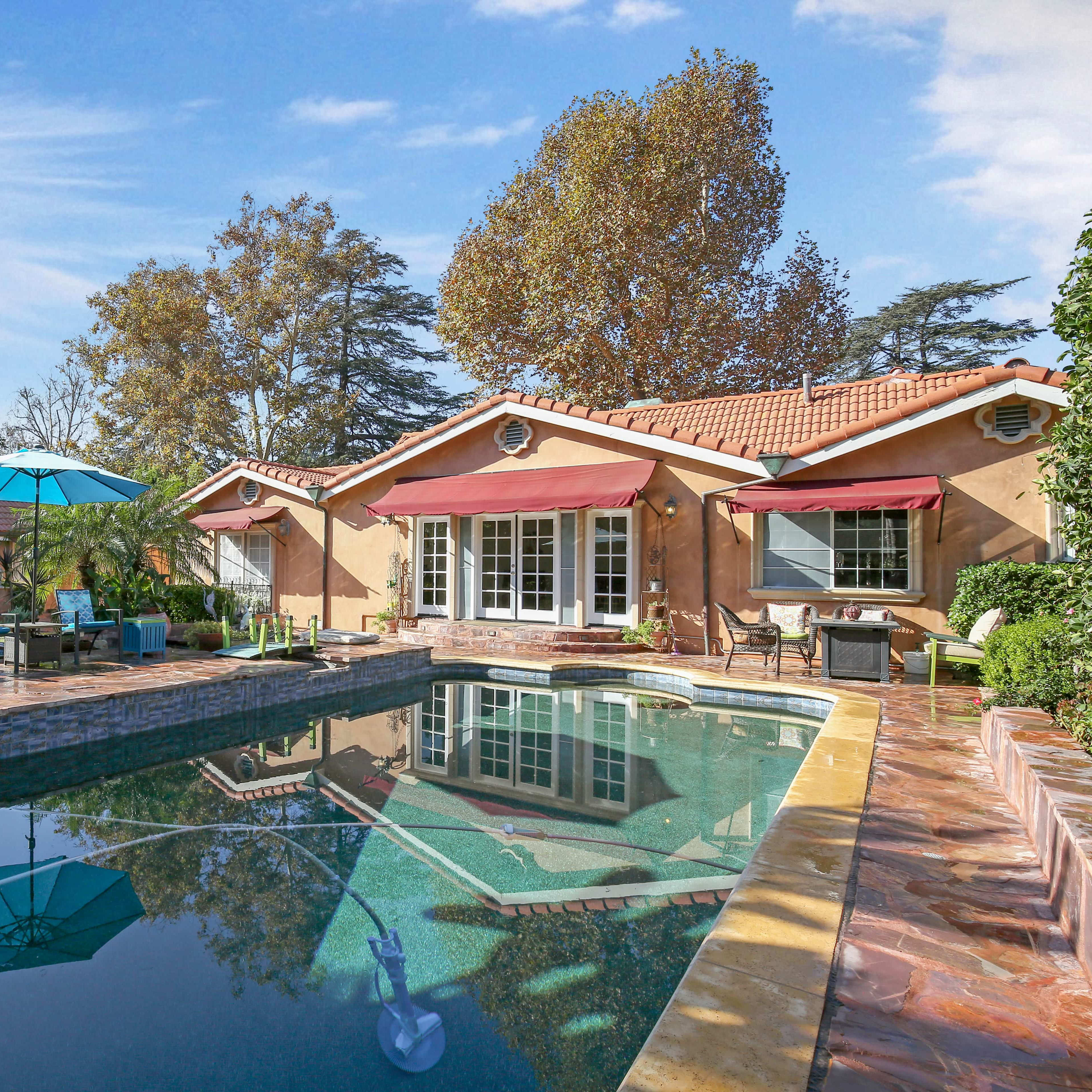 A backyard with a swimming pool surrounded by lounge chairs and an umbrella, adjacent to a house with a landscaped garden.
