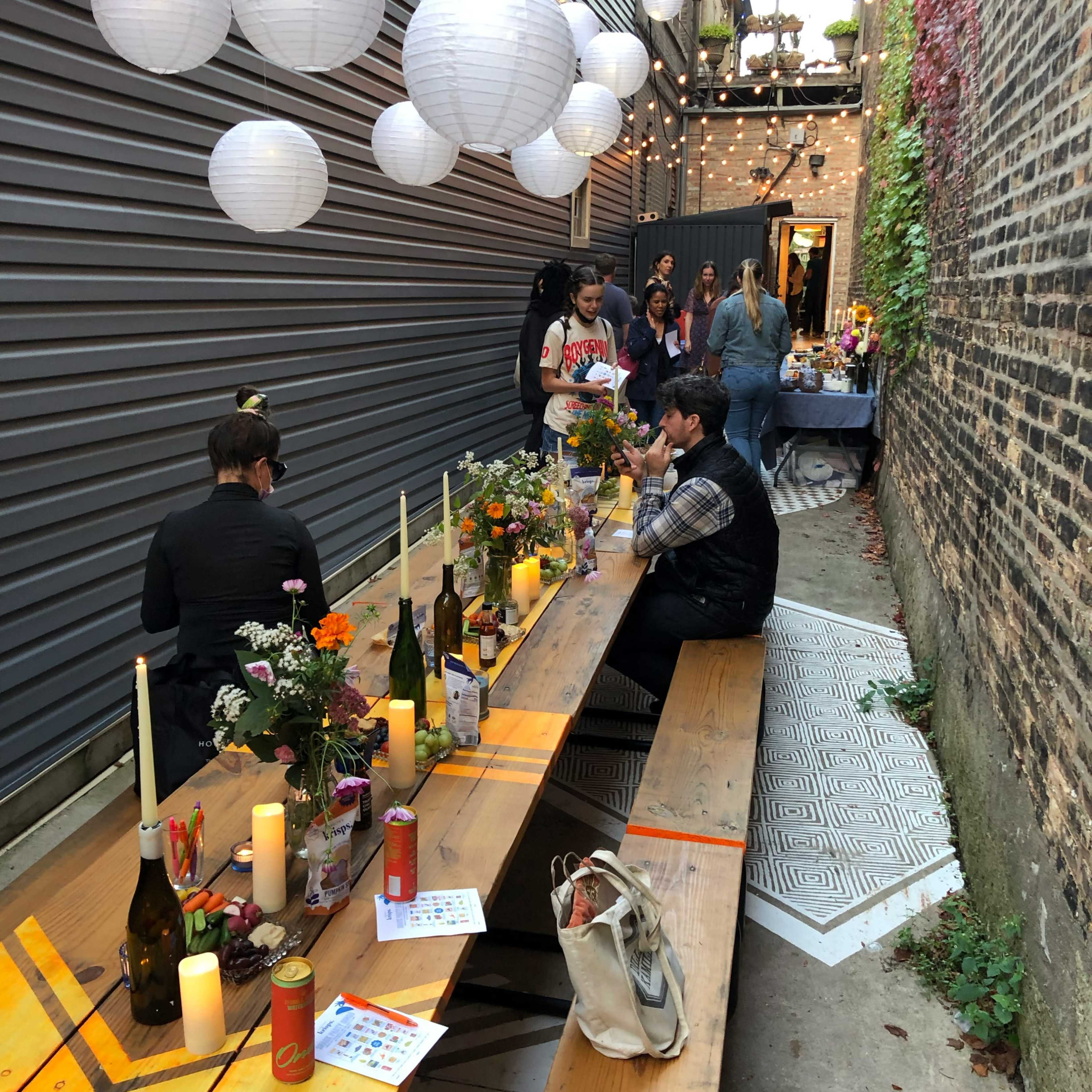 A narrow alley decorated with string lights and paper lanterns, featuring a long wooden table set for a gathering with candles and flowers.