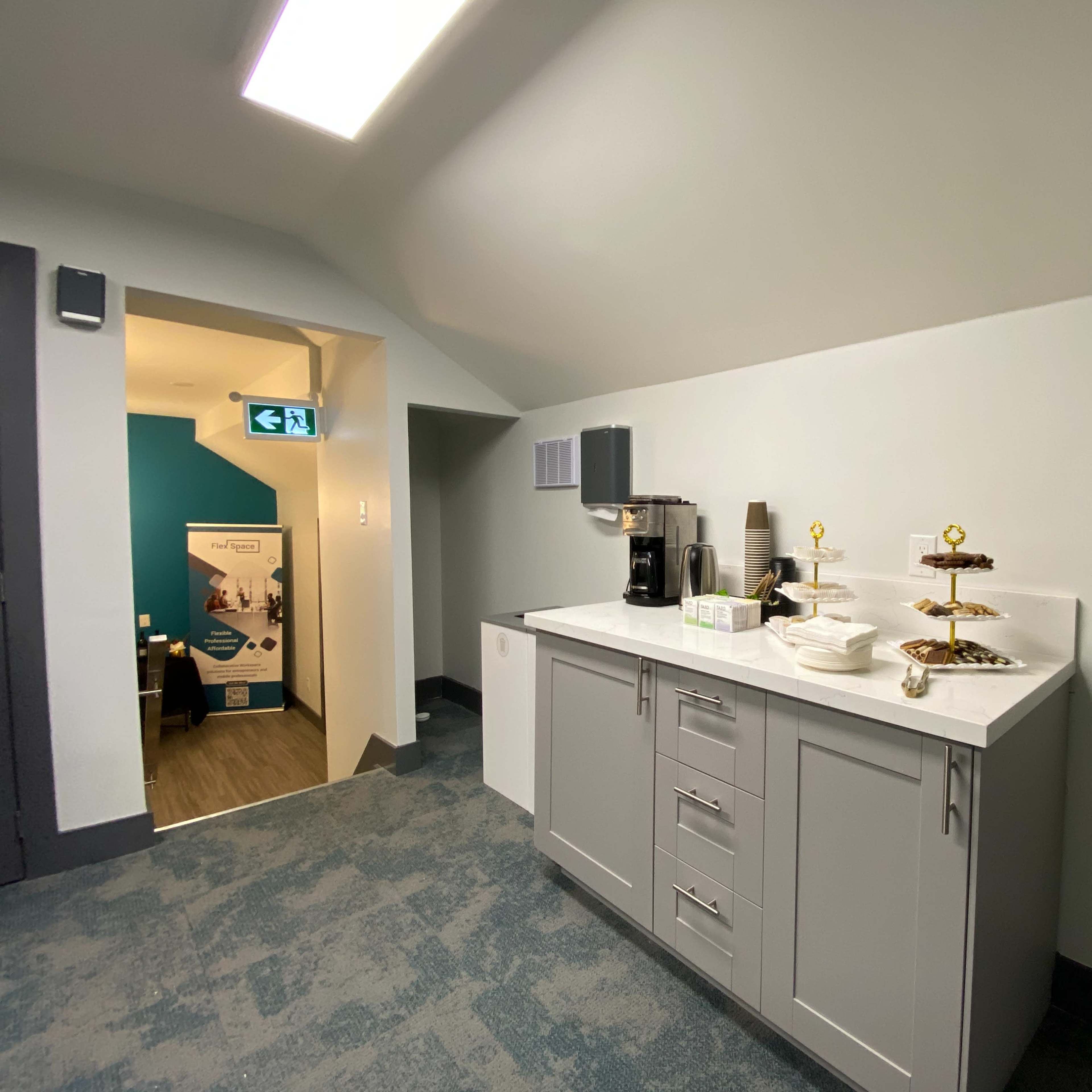A modern kitchenette with a countertop, coffee maker, and a tiered display of pastries, located near a doorway leading to a blue-walled room.