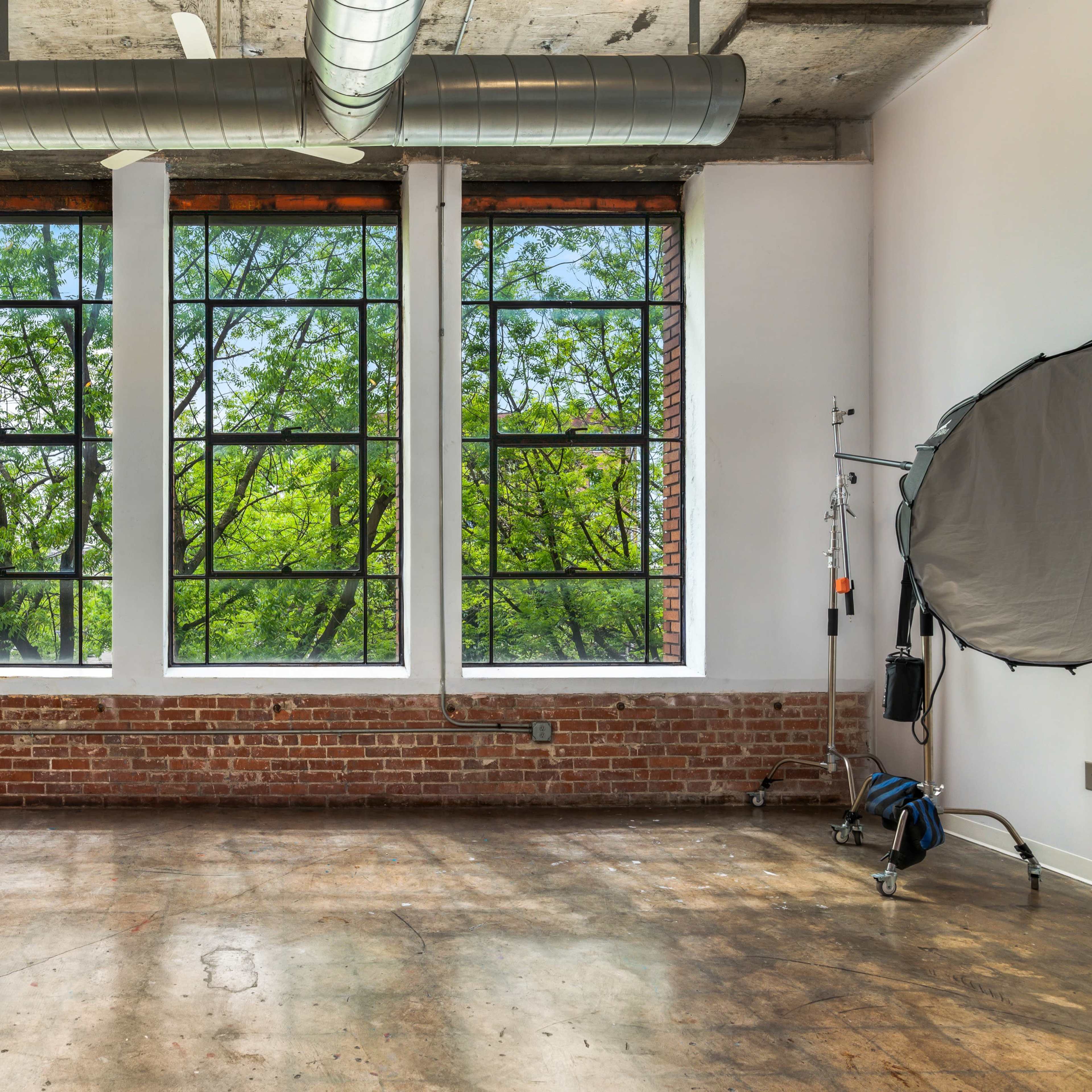 A spacious room with large windows overlooking green trees, featuring exposed brick walls and concrete flooring.