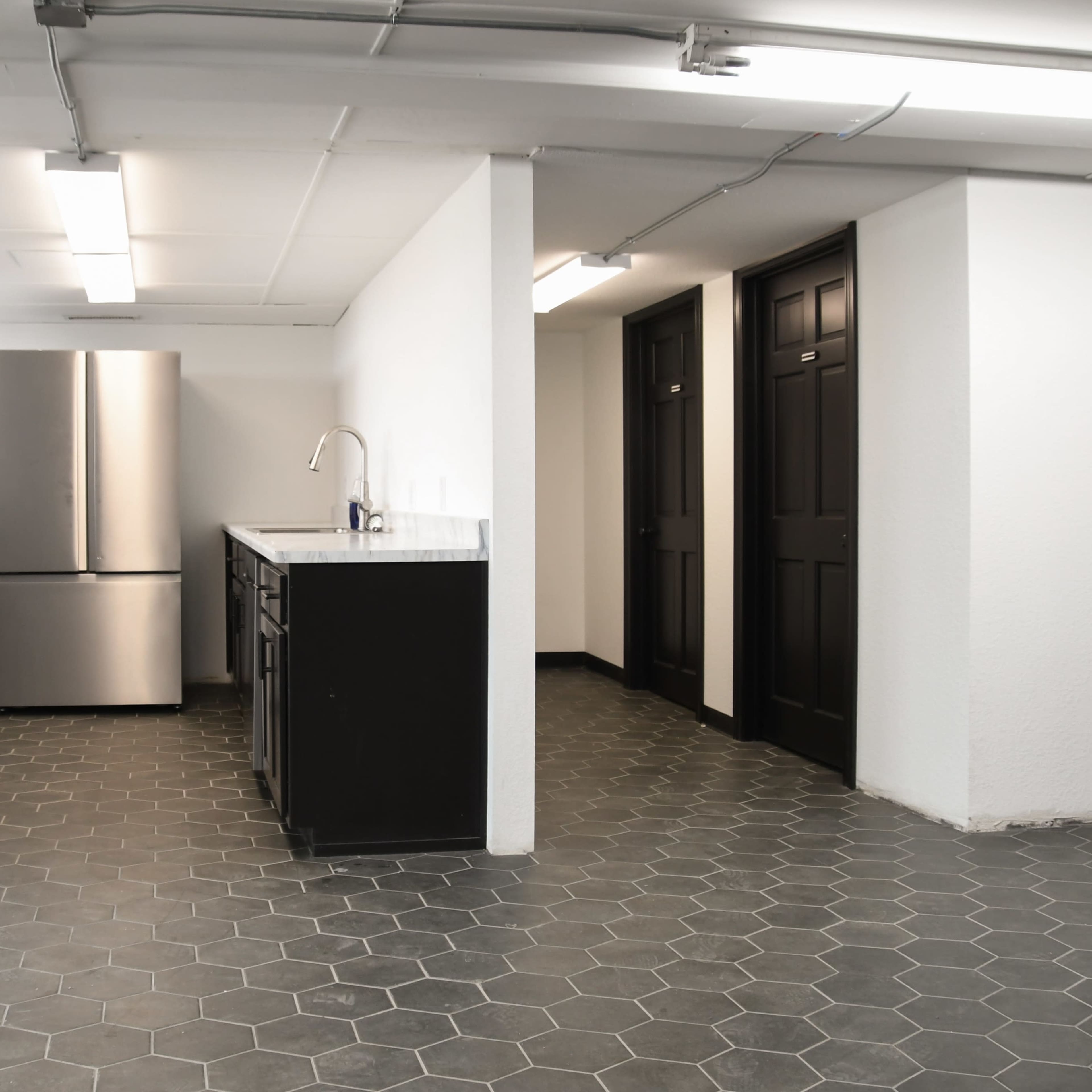 A spacious room features a stainless steel refrigerator, dark counters, and two closed doors along one wall, with a hexagonal tile floor.