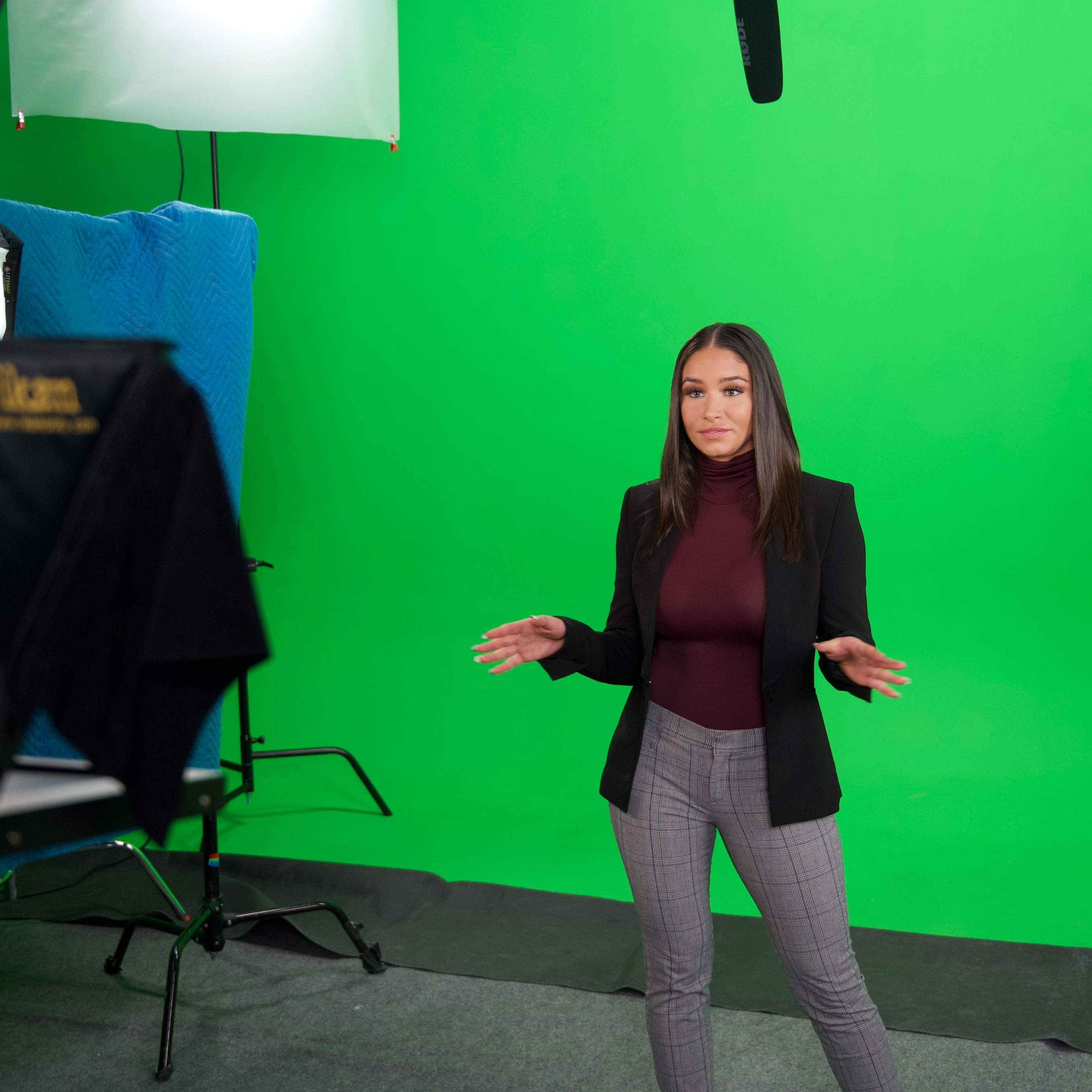 A woman stands in front of a green screen, gesturing with her hands while preparing for a presentation, with studio lighting and equipment visible.