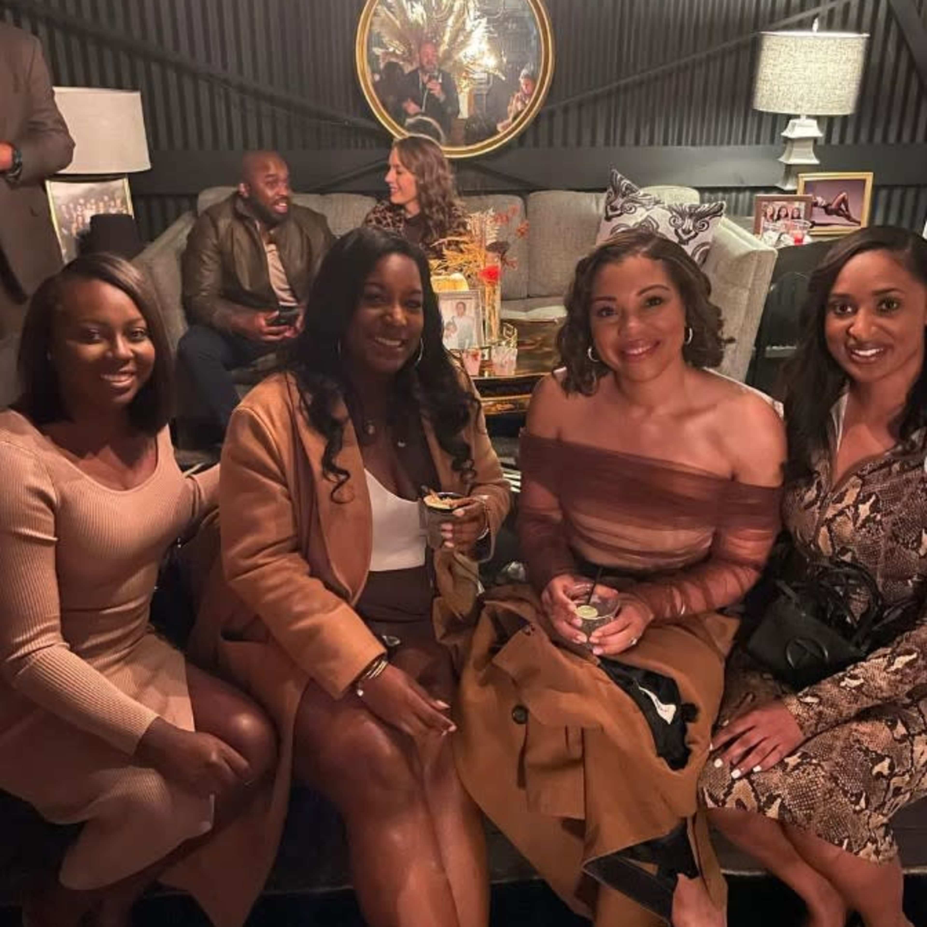 Four women are posing for a photo while seated in a cozy lounge area, with two people visible in the background engaged in conversation.