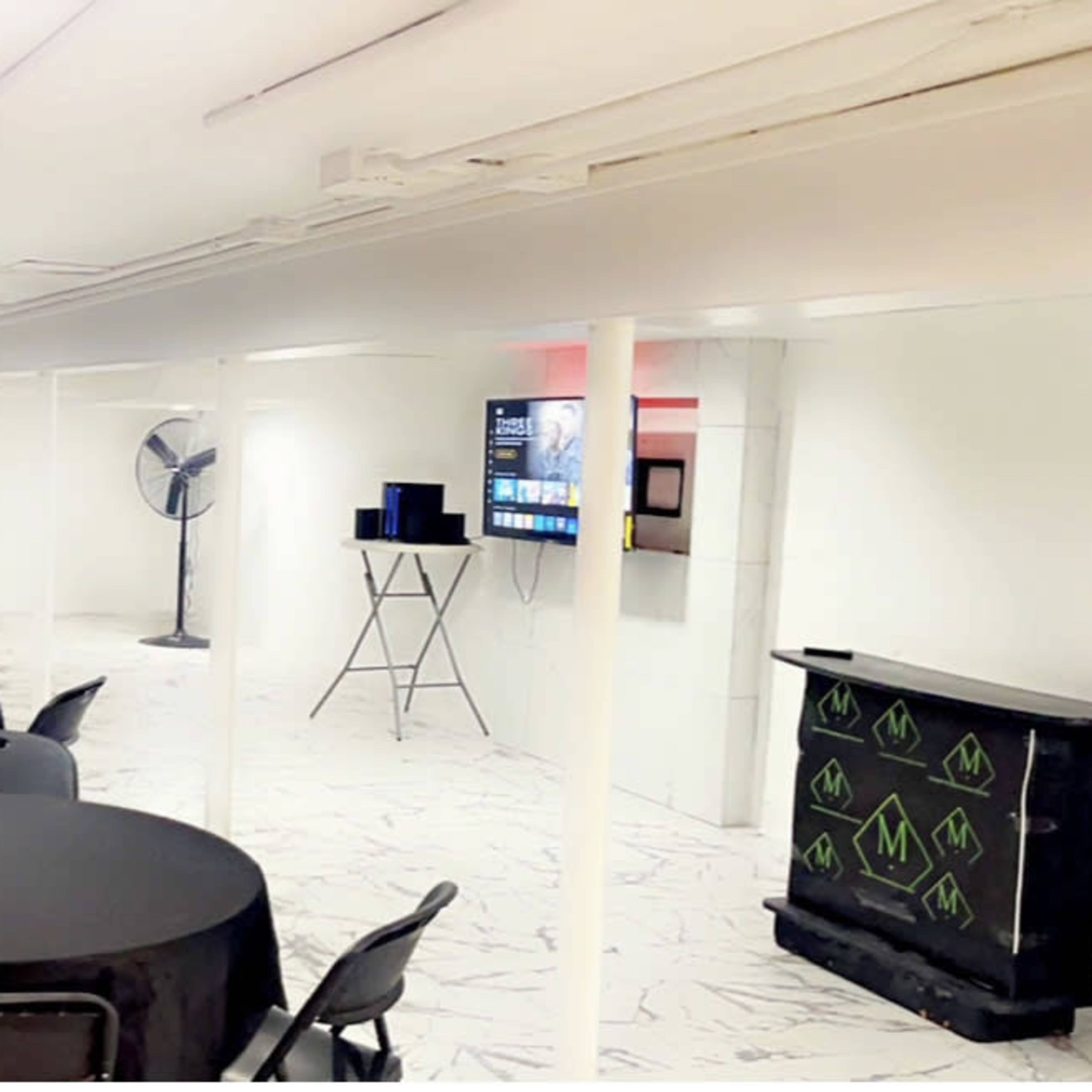 A brightly lit, minimalist event space with black tables and chairs arranged throughout, featuring a television mounted on the wall and a fan in the corner.