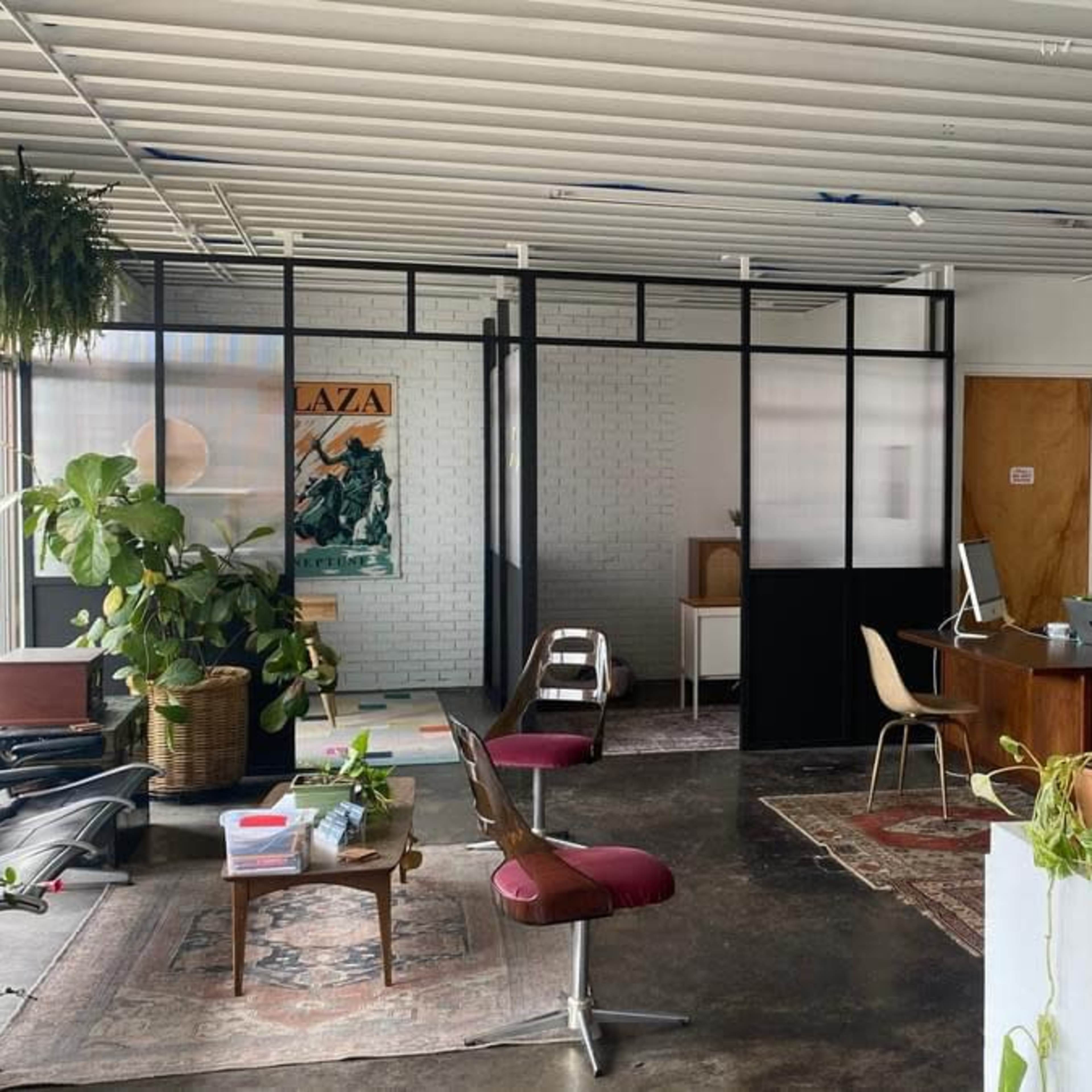 A modern office space with a combination of plants, furniture, and workstations, featuring a glass partition separating different areas.