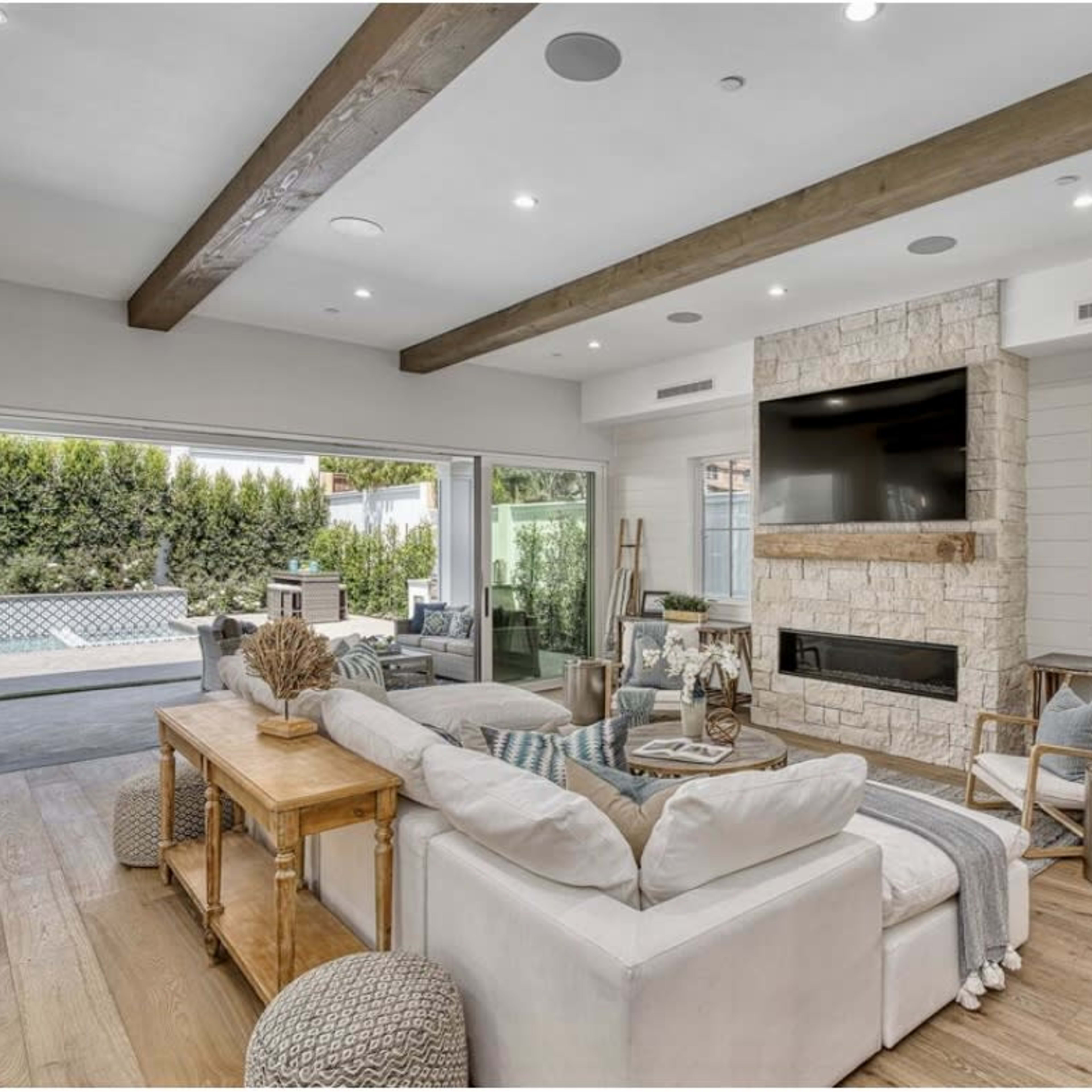 The living room features a light-colored sectional sofa, a stone fireplace, and large windows opening to an outdoor space with a pool.