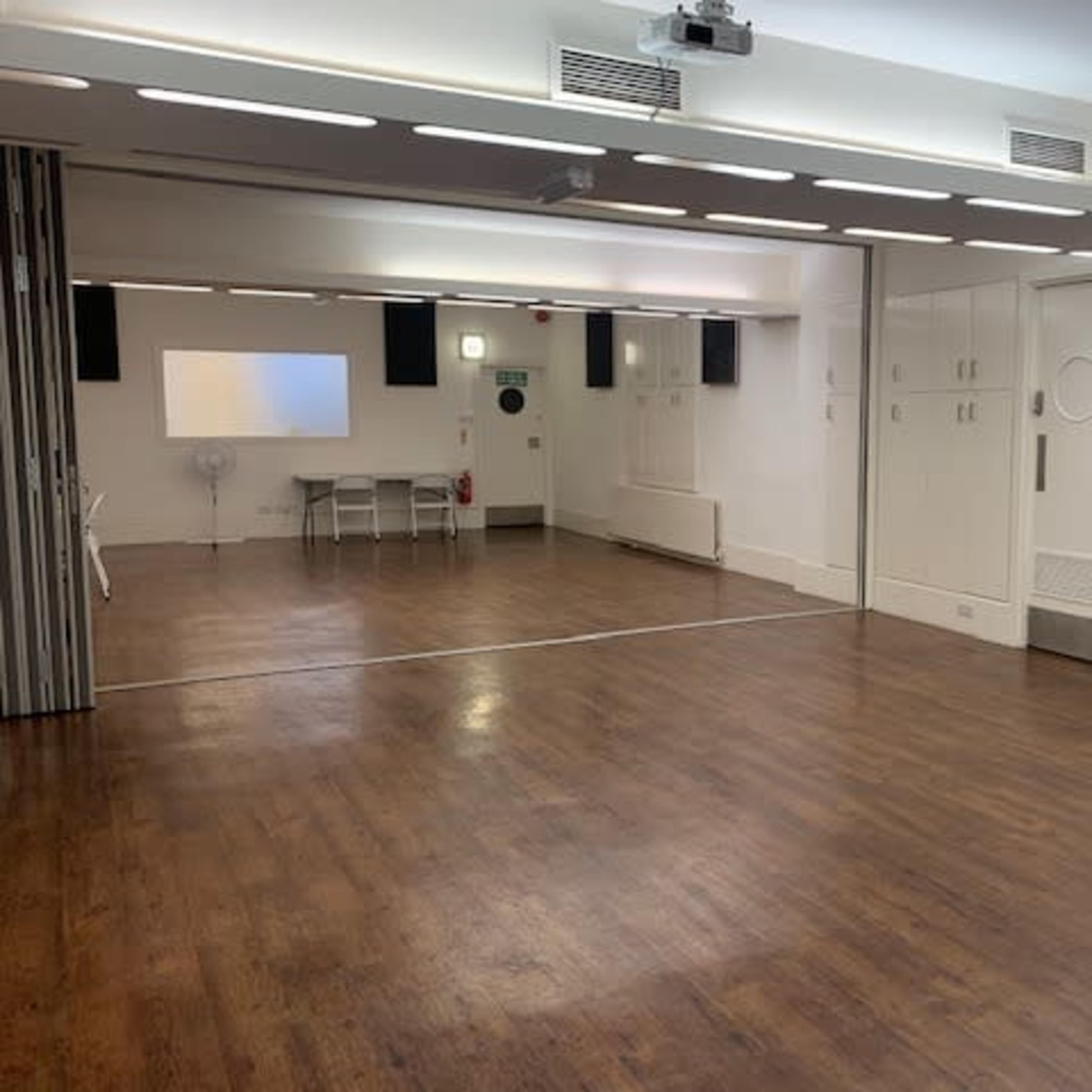 A spacious, modern room features wooden flooring, white walls, and a projection screen at one end, with a table and chairs set up in the center.