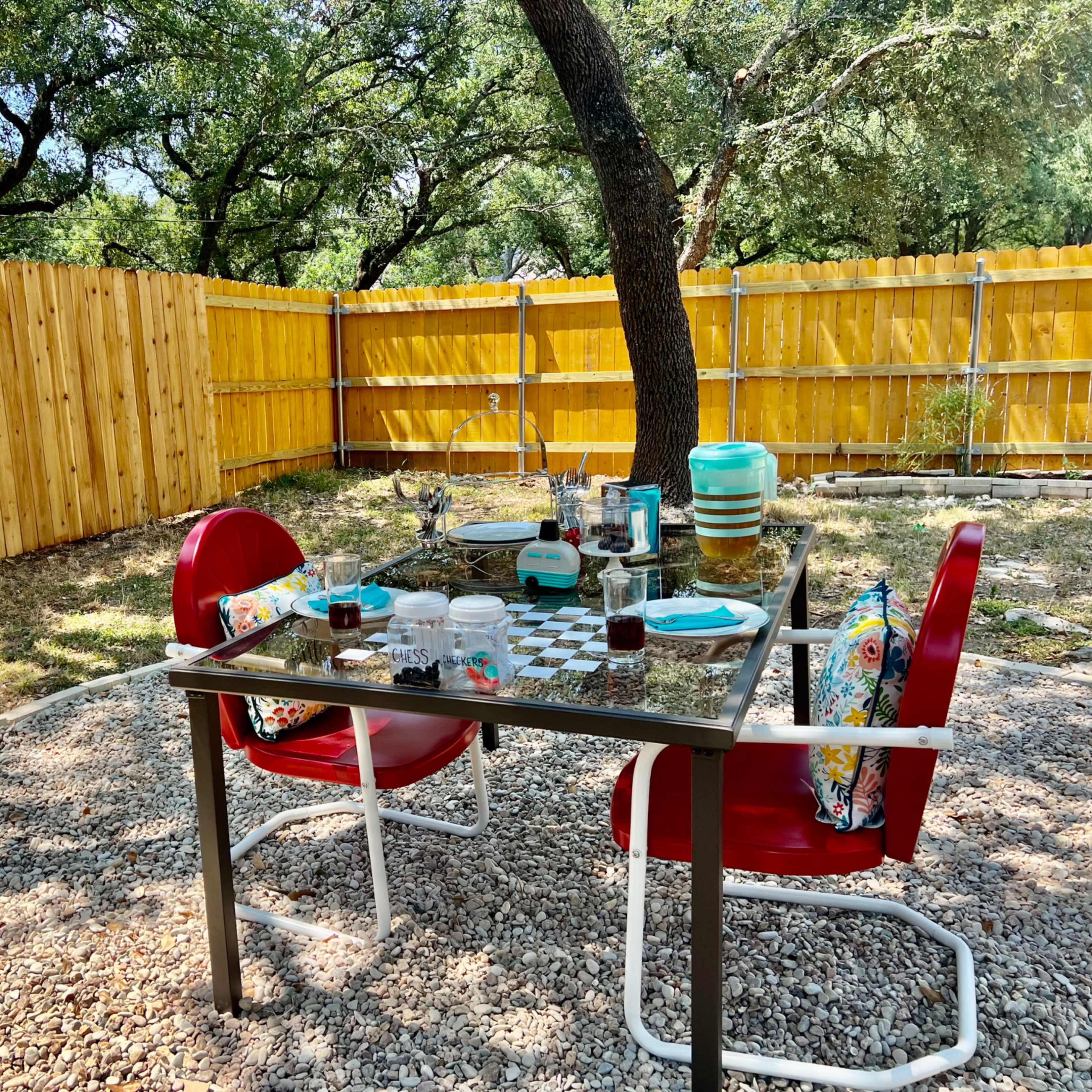 A table set with dinnerware and drinks is surrounded by two red chairs in a yard enclosed by a wooden fence.