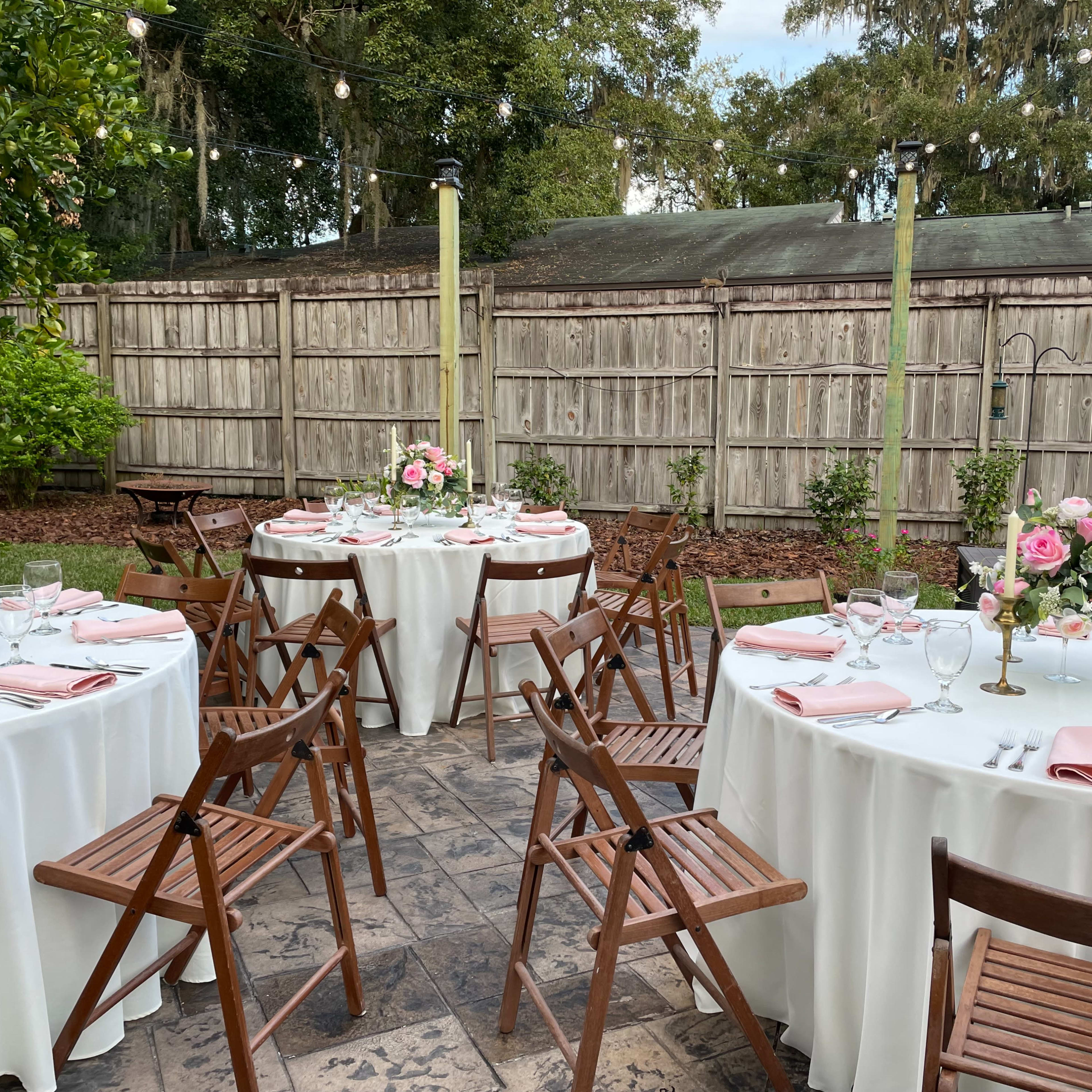 A decorated outdoor dining area with round tables covered in white tablecloths and set with pink napkins, surrounded by wooden chairs and floral centerpieces.