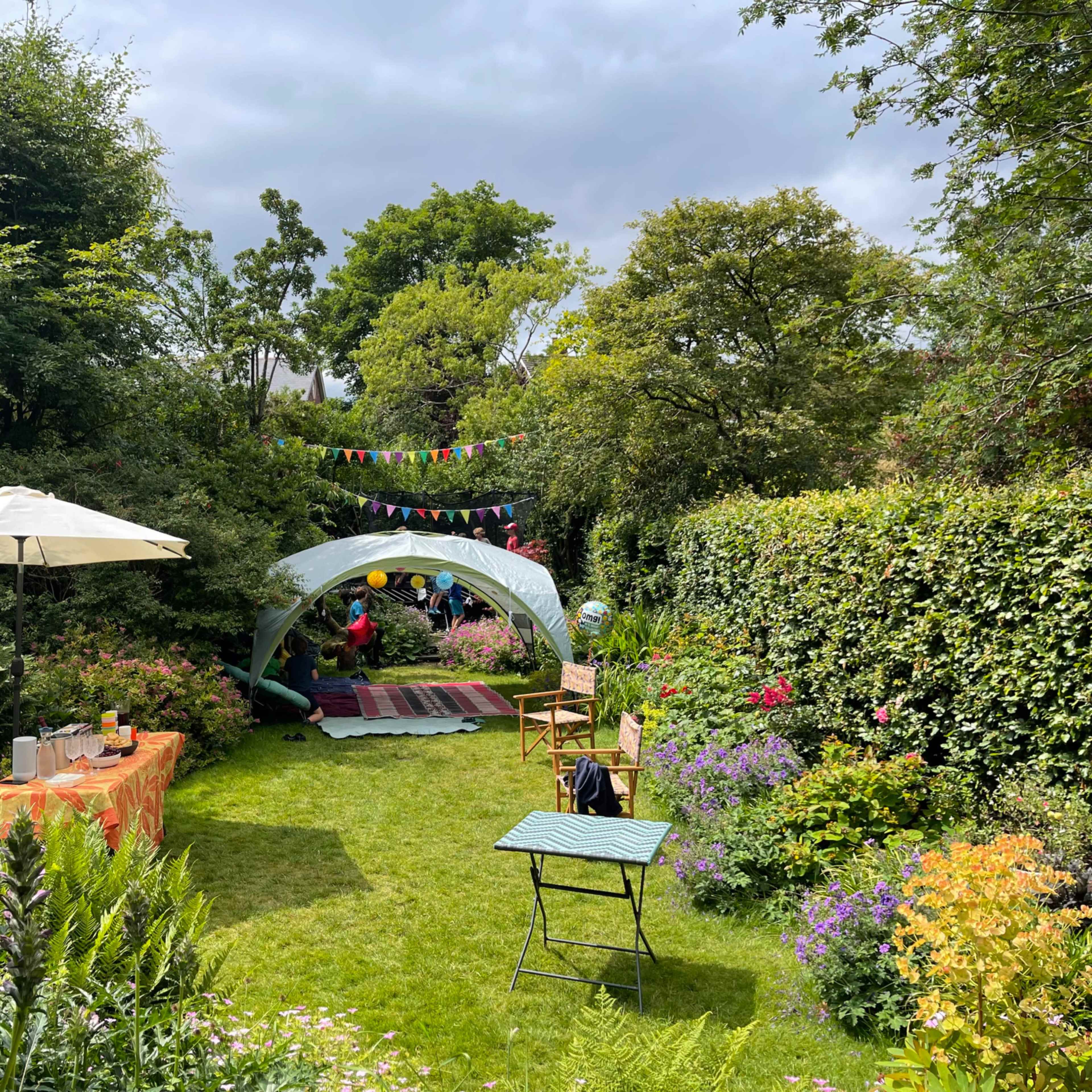A garden setting features a decorated tent, picnic tables, and colorful bunting amid lush greenery and blooming flowers.