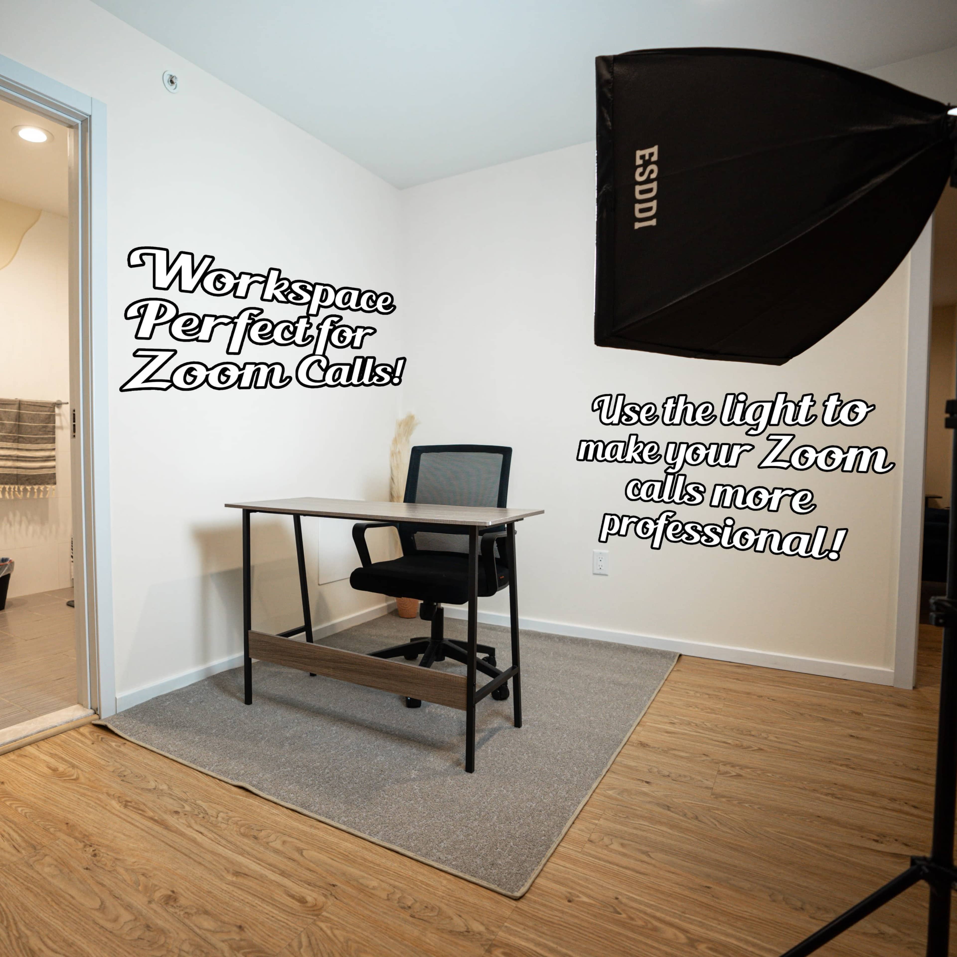 A small modern workspace featuring a desk and chair in a room, with a bathroom doorway visible in the background and a softbox light set up for video calls.