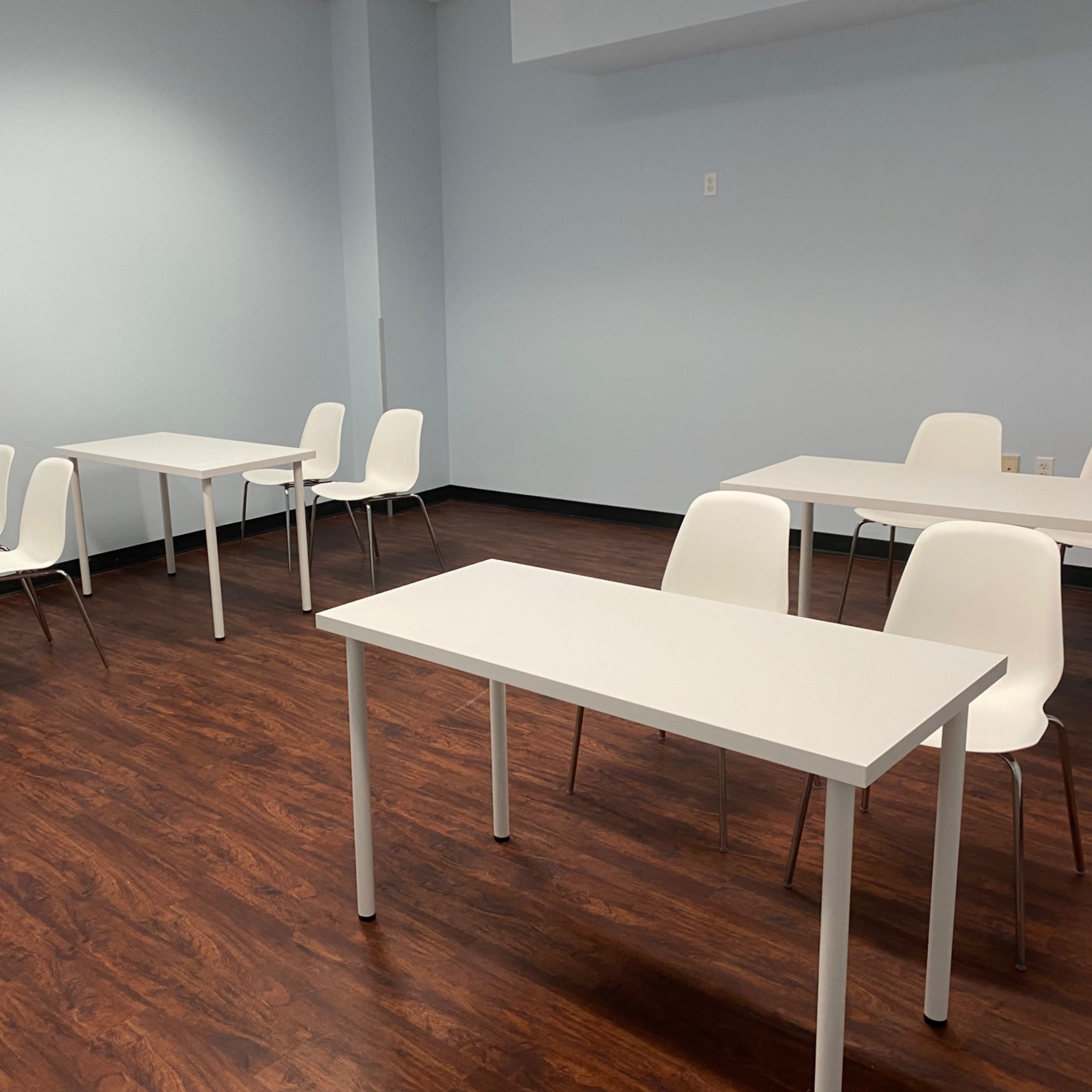 A minimalistic classroom features several white tables and chairs arranged in a spacious, well-lit area with wooden flooring.