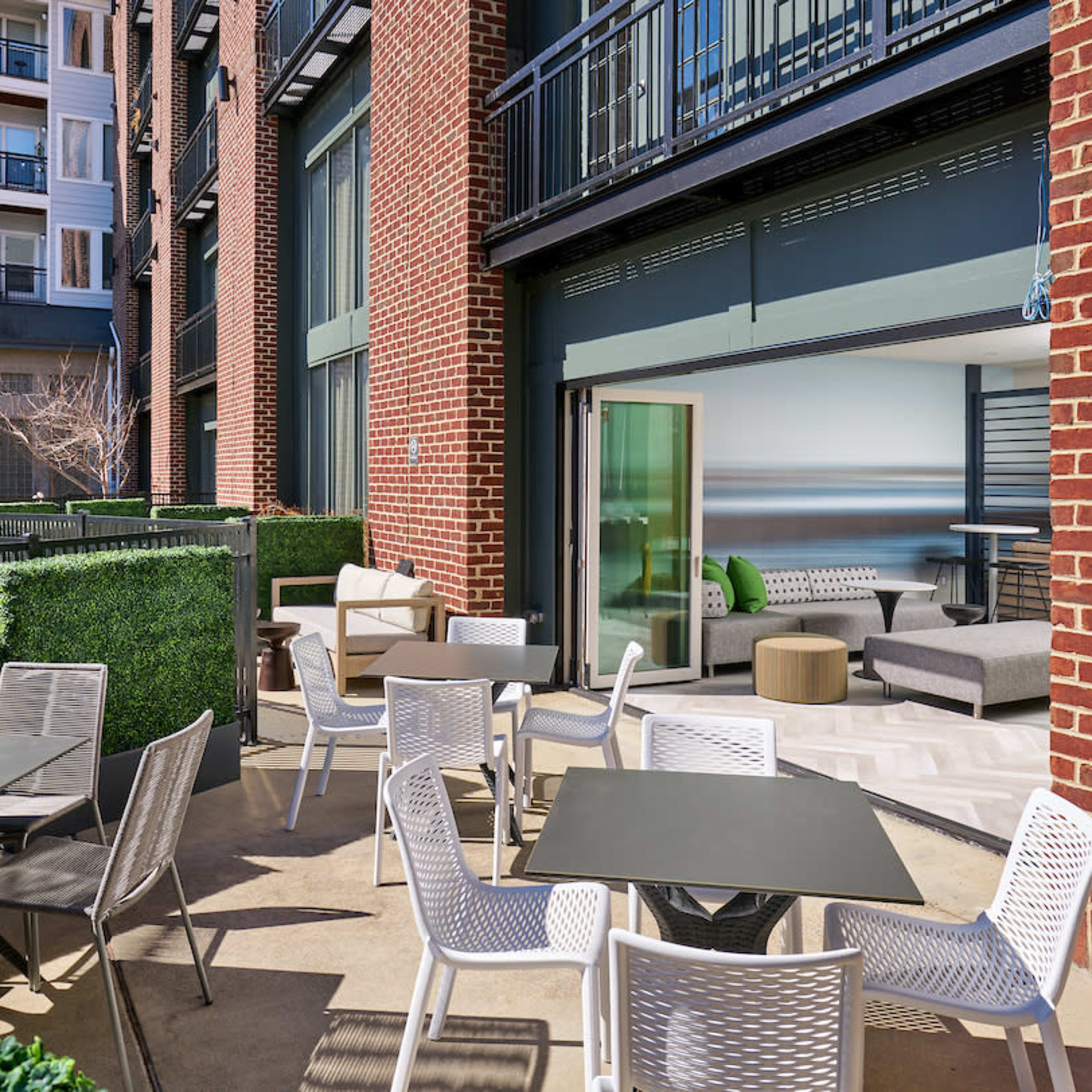 An outdoor patio area with tables and chairs, surrounded by greenery, next to a building with large sliding glass doors.