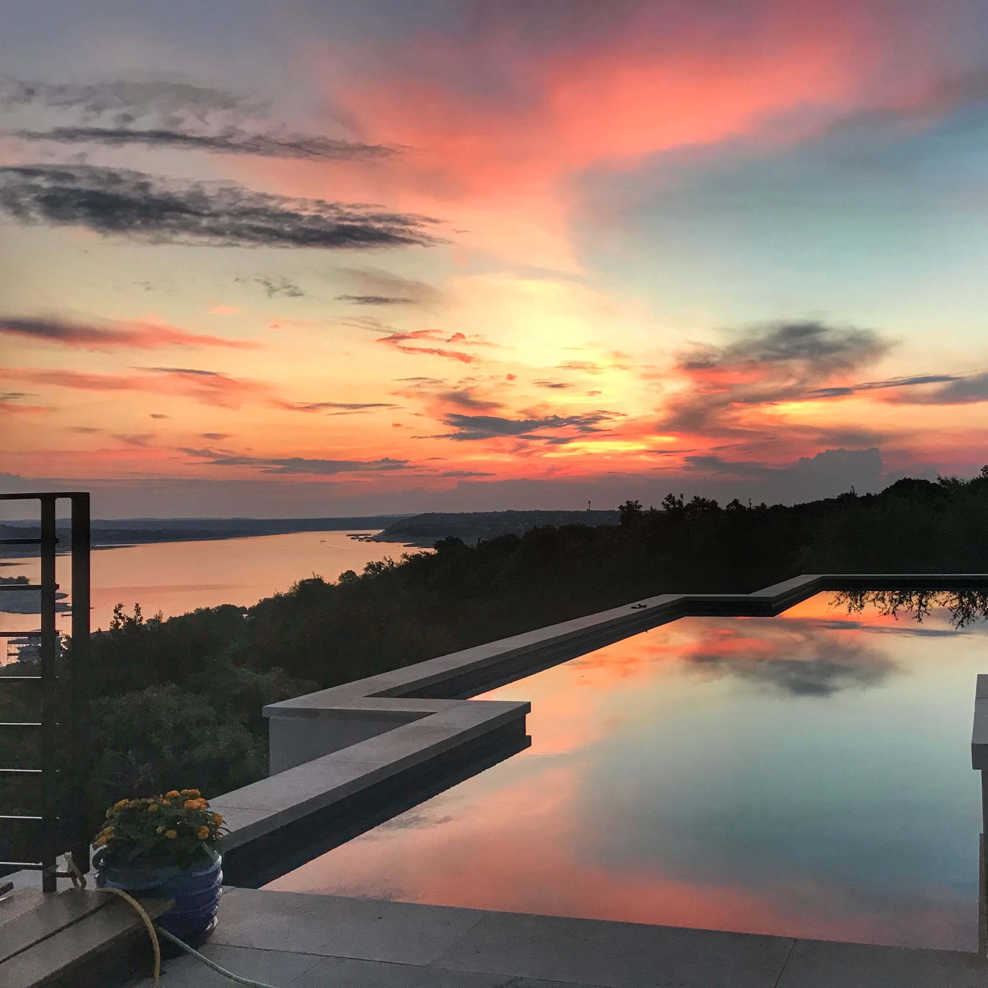 A tranquil infinity pool reflecting a colorful sunset over a lake and surrounding landscape.