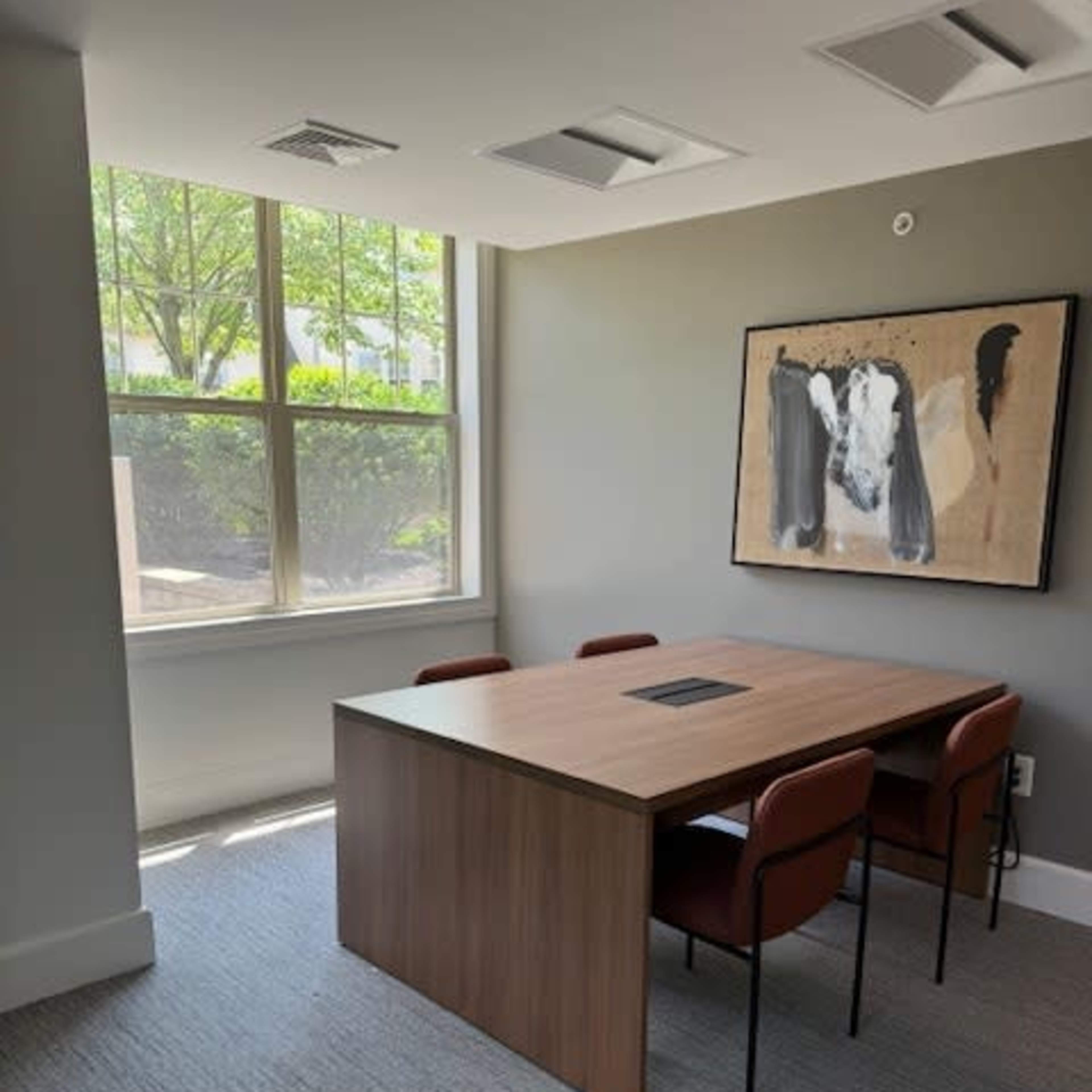 A small conference room featuring a wooden table surrounded by four chairs, with large windows letting in natural light and a piece of art on the wall.