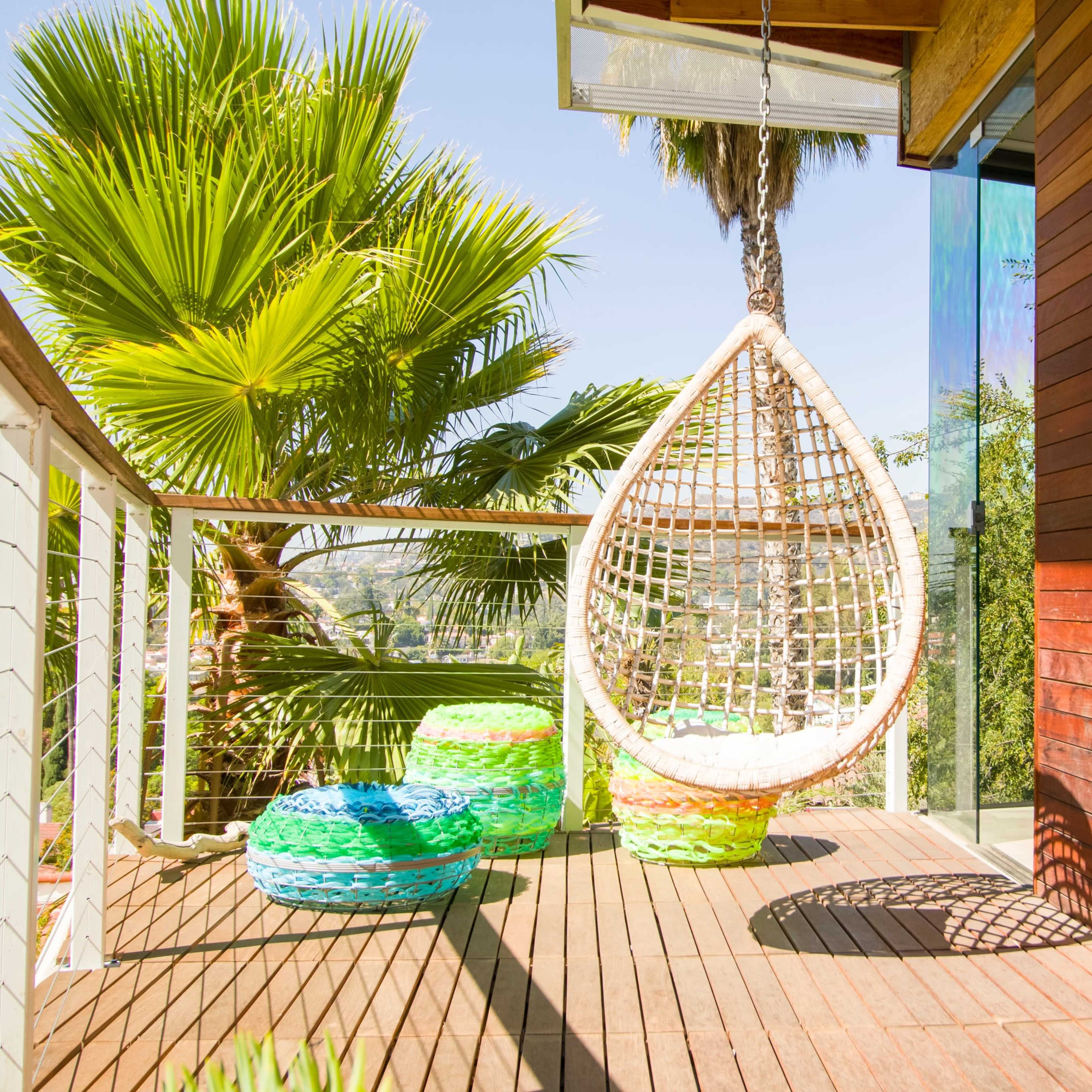 A hanging wicker chair is positioned on a wooden deck beside colorful poufs and surrounded by lush palm trees.