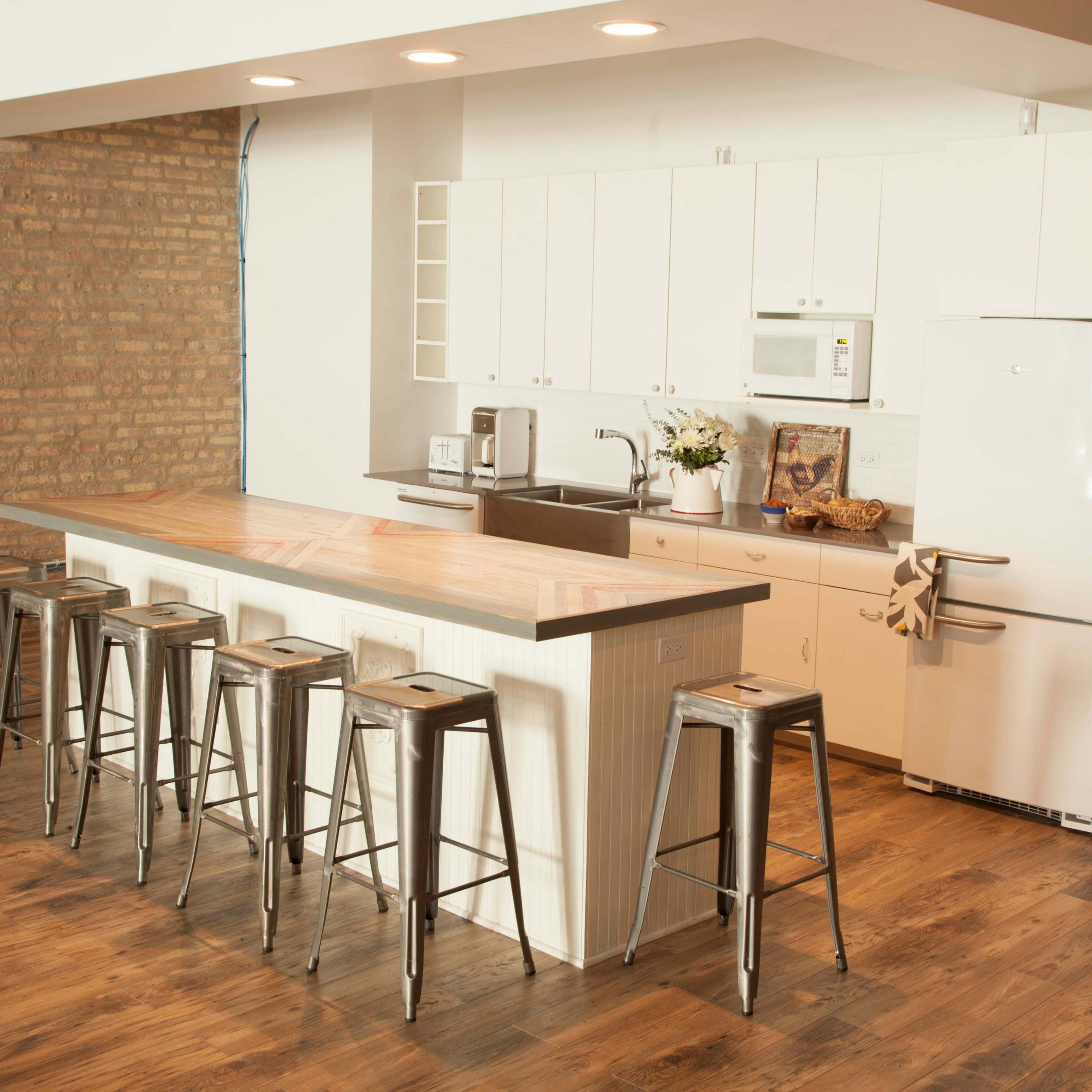 A modern kitchen featuring a long wooden island with metal bar stools, white cabinetry, a refrigerator, and exposed brick walls.
