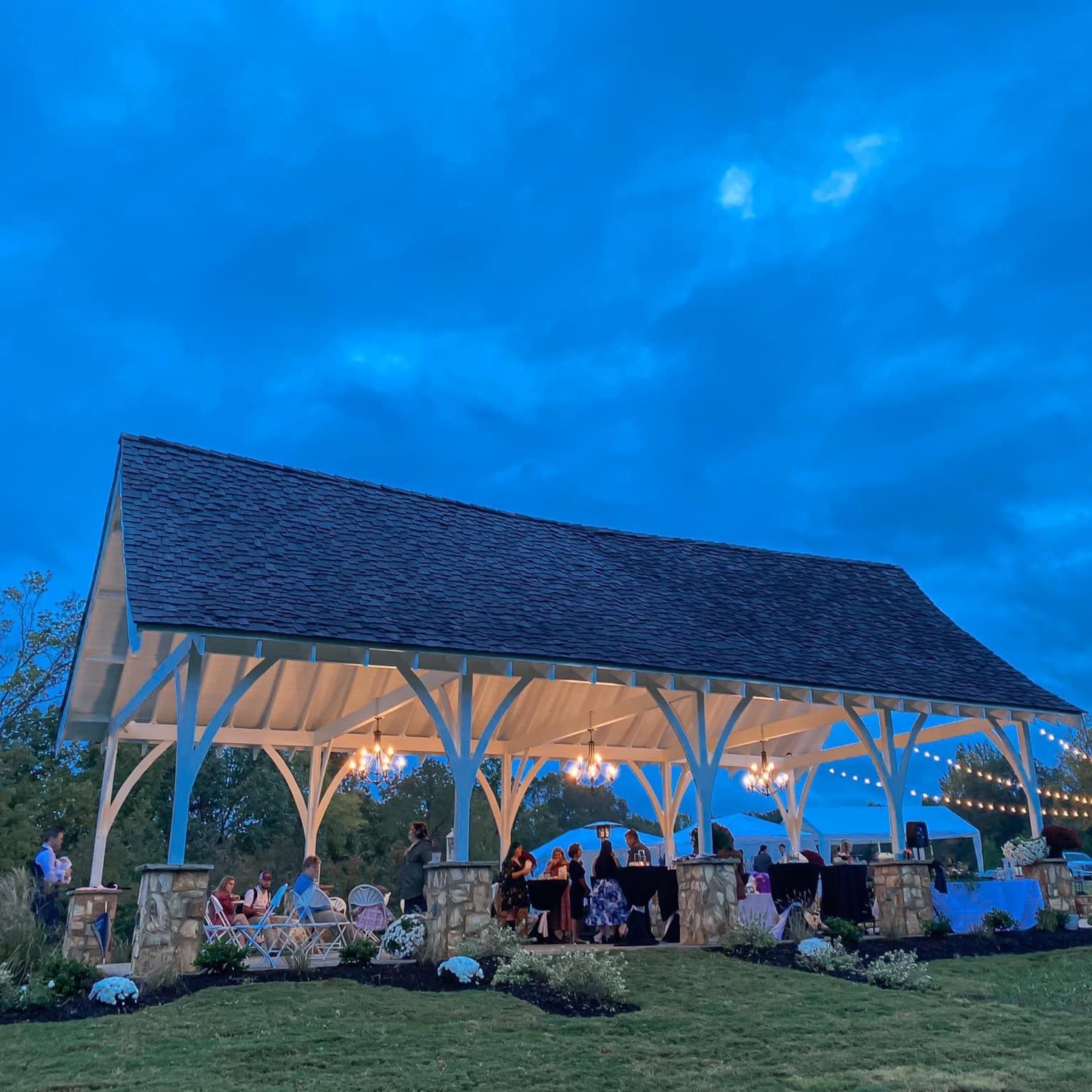 A covered pavilion with string lights and a gathering of people is set against a twilight sky.