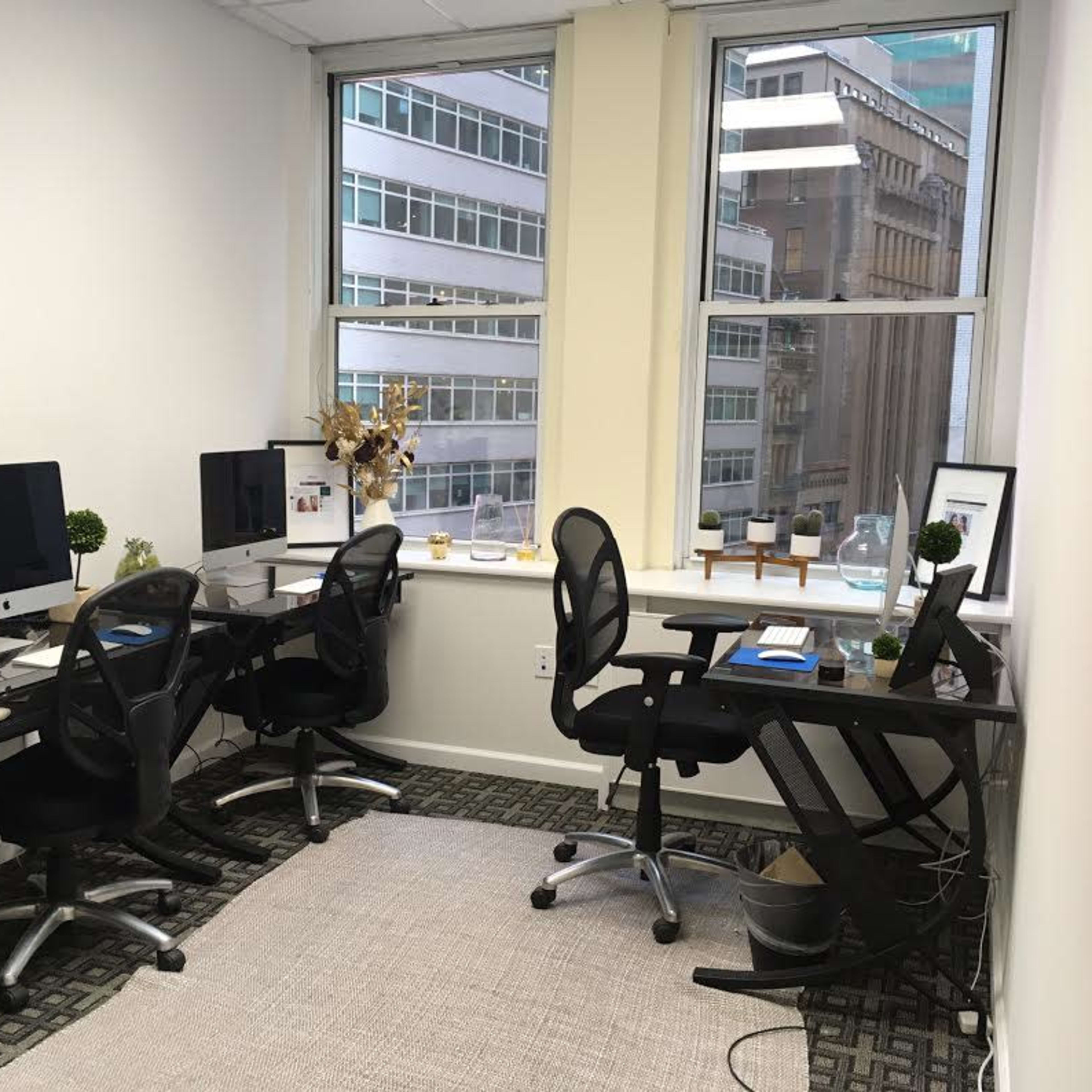 A bright office space with four workstations equipped with computers and ergonomic chairs, alongside large windows overlooking a cityscape.