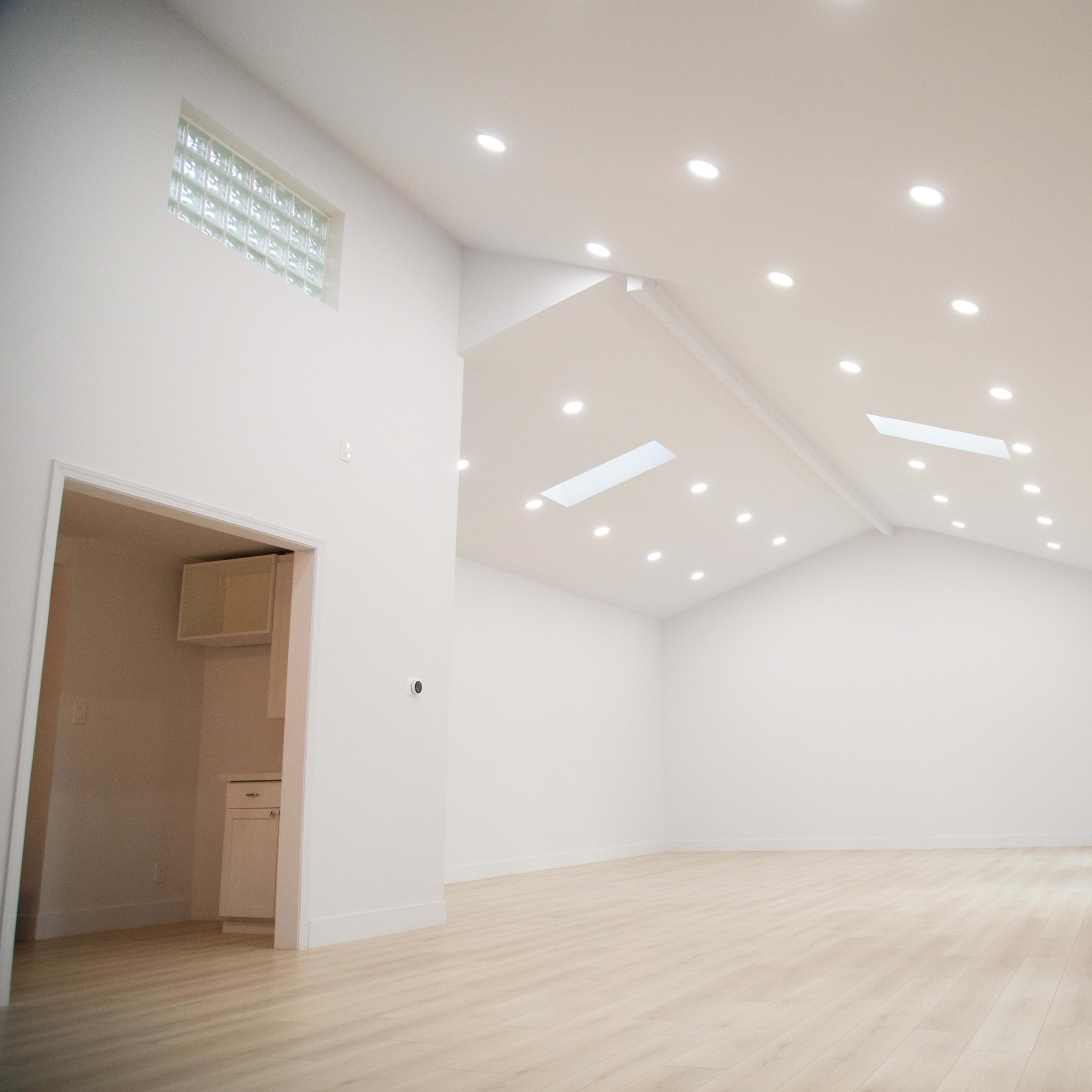 A spacious interior room features a high ceiling with recessed lighting and white walls, complemented by large windows and a light wooden floor.